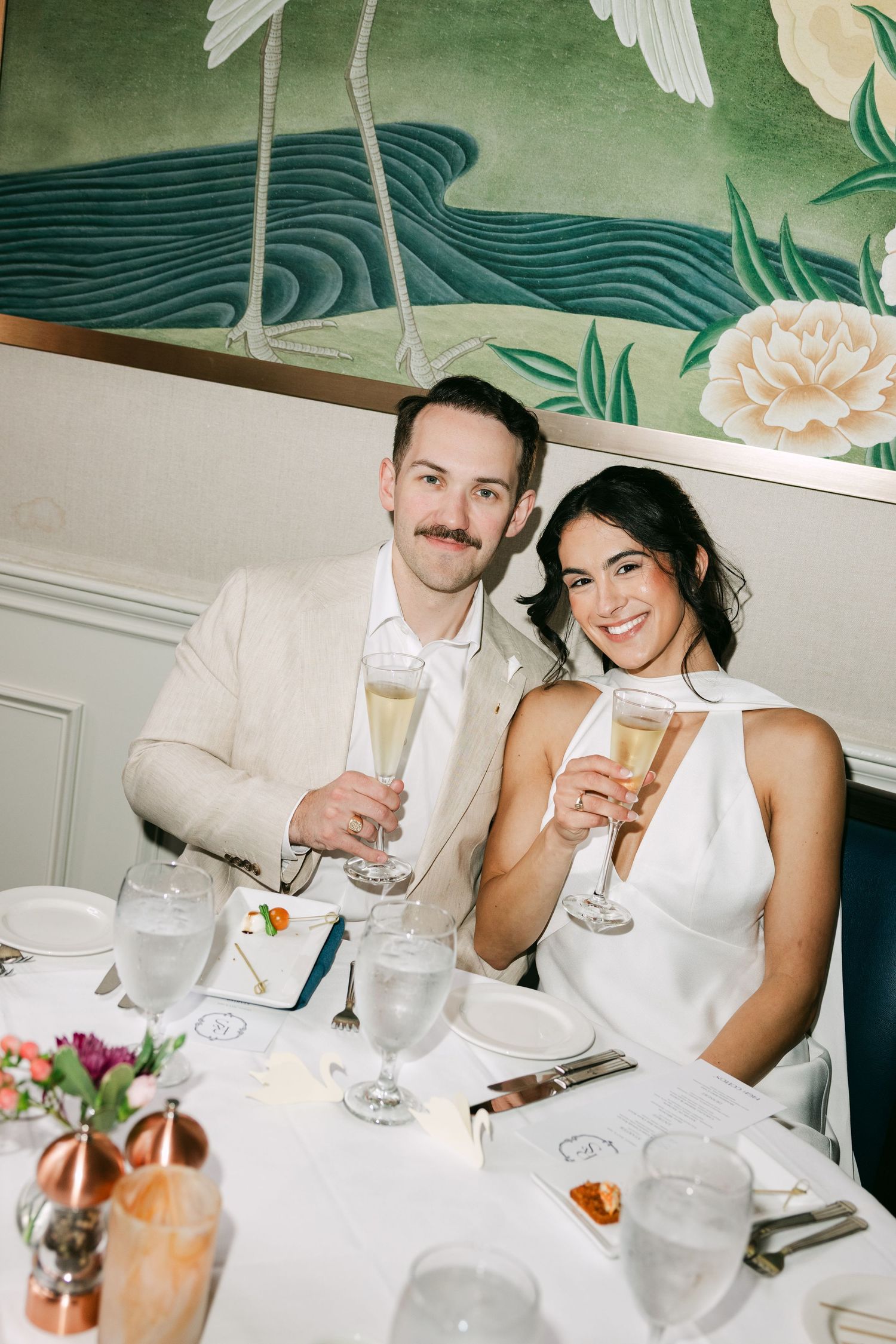 Smiling bride and groom toast with champagne glasses at their elegant wedding reception dinner table.