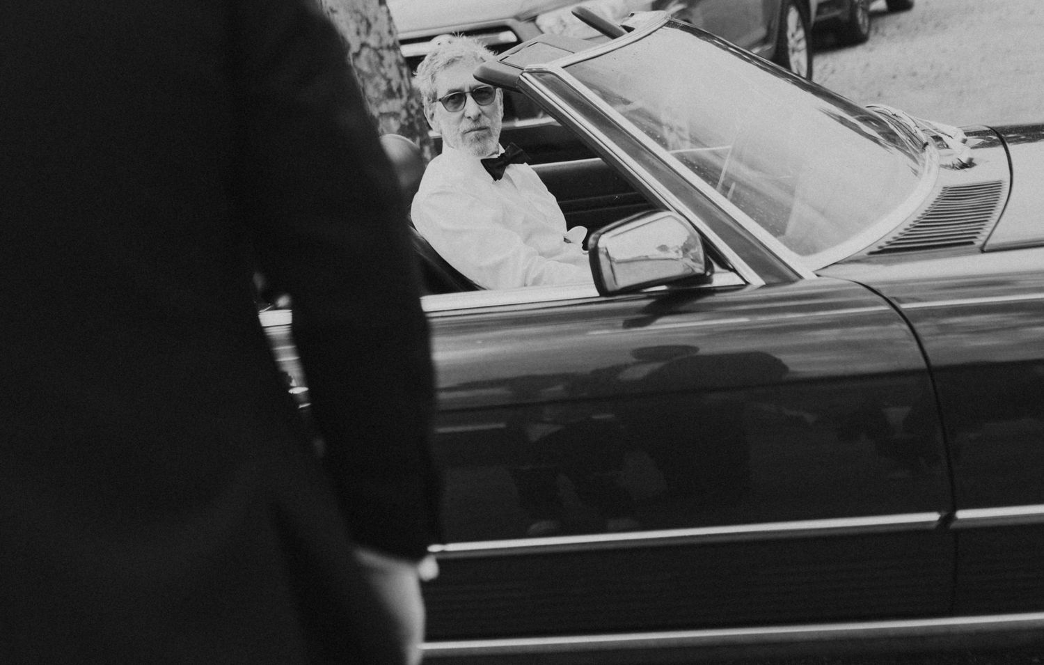 Black and white photo of an elderly person sitting in a classic convertible car, viewed from outside the vehicle.