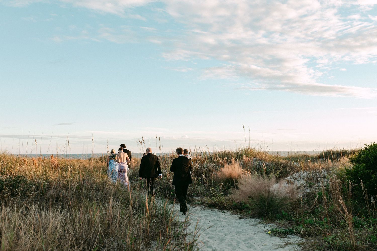 Wedding party walks sandy path through sea grass toward ocean at sunset beach wedding.