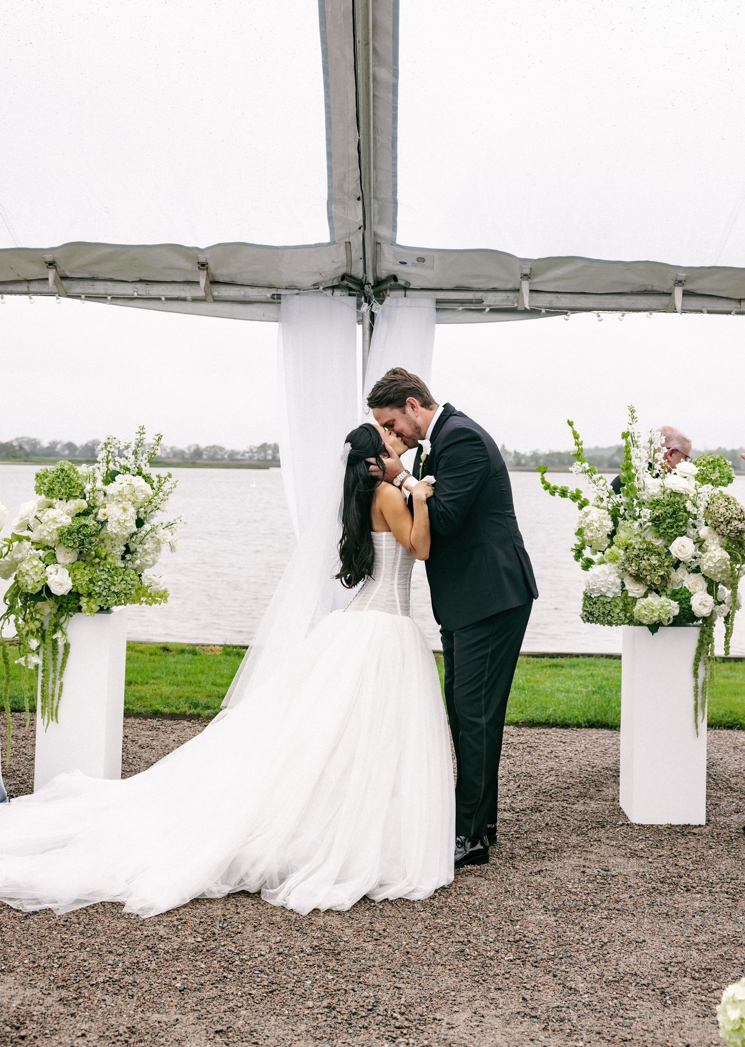 Bride and groom share first kiss under tent altar with white floral arrangements by waterfront.