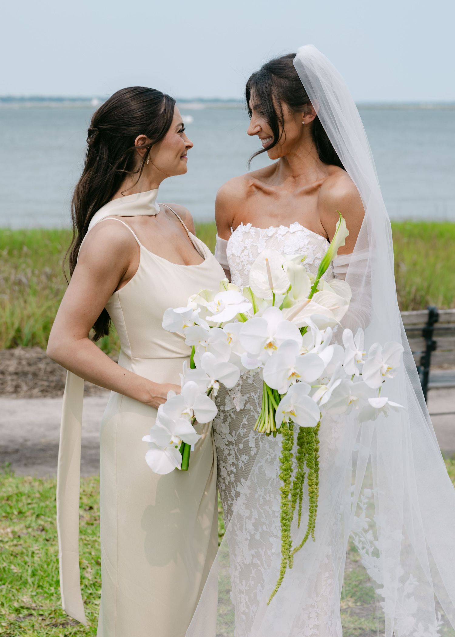 A bride and bridesmaid share an intimate moment at a seaside wedding with white orchids and a flowing lace veil.