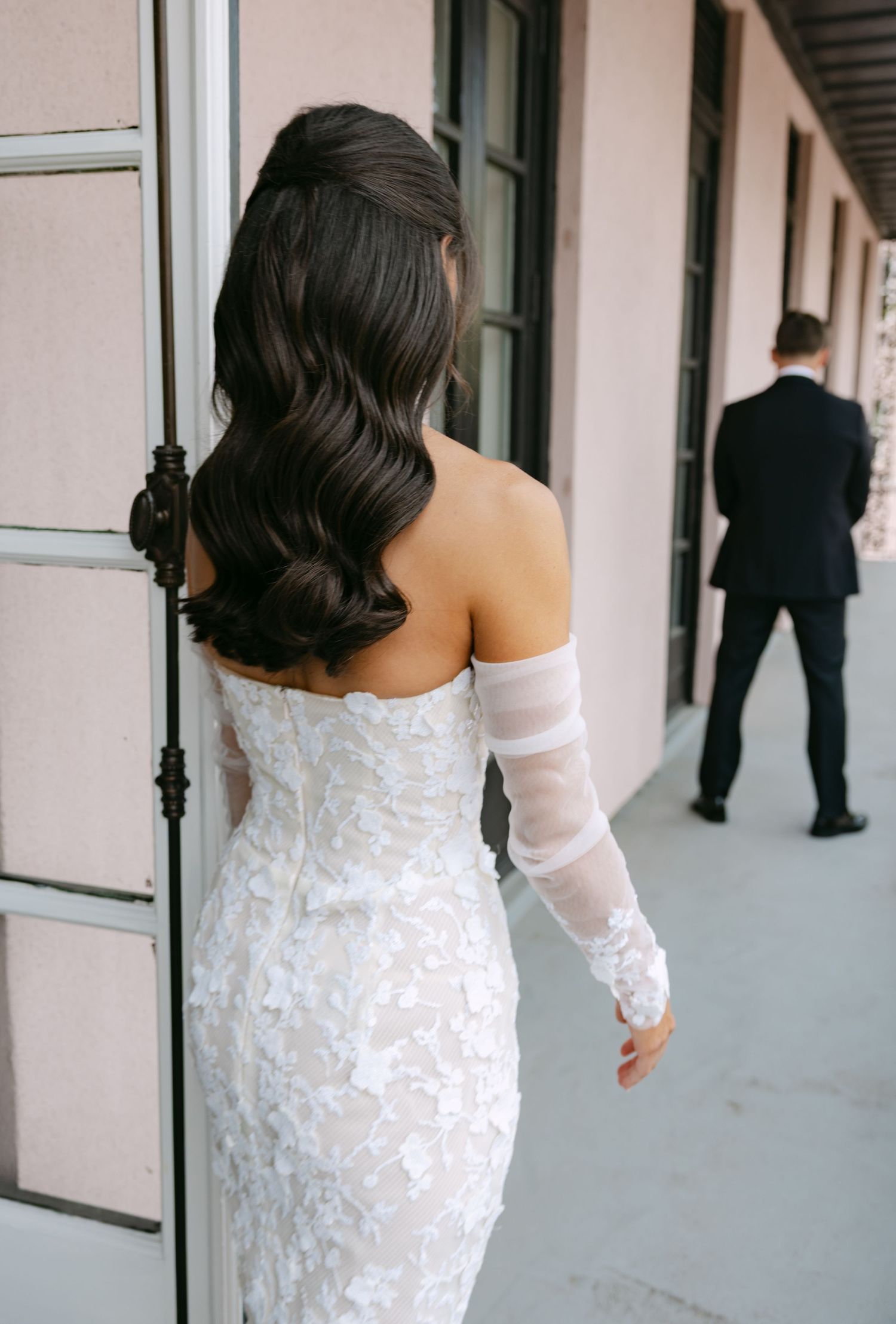 A bride in a fitted lace gown and sheer sleeves stands in a hallway while a groom waits in the distance.