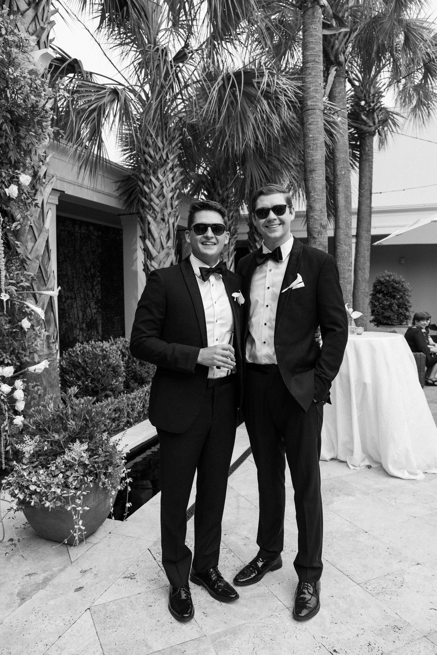 Two people in formal black tuxedos pose together on a paved walkway surrounded by palm trees.