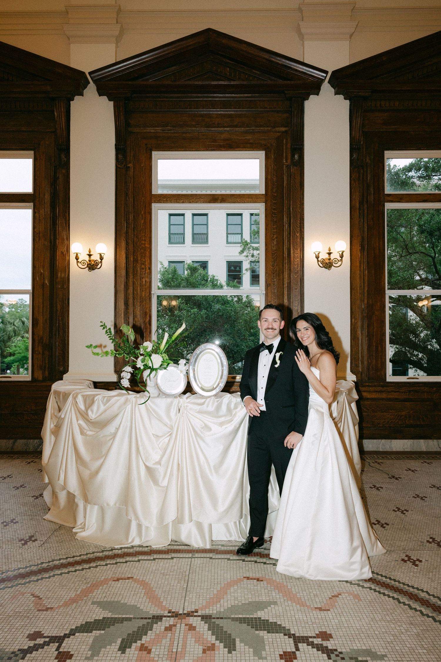 Bride and groom posing together in elegant wedding attire beside a draped table with floral arrangement in a grand venue.