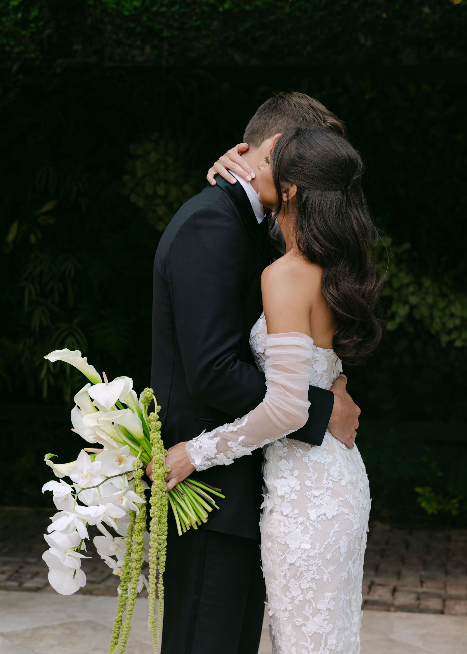 Romantic wedding couple embraces by lush greenery while holding a white orchid bouquet.