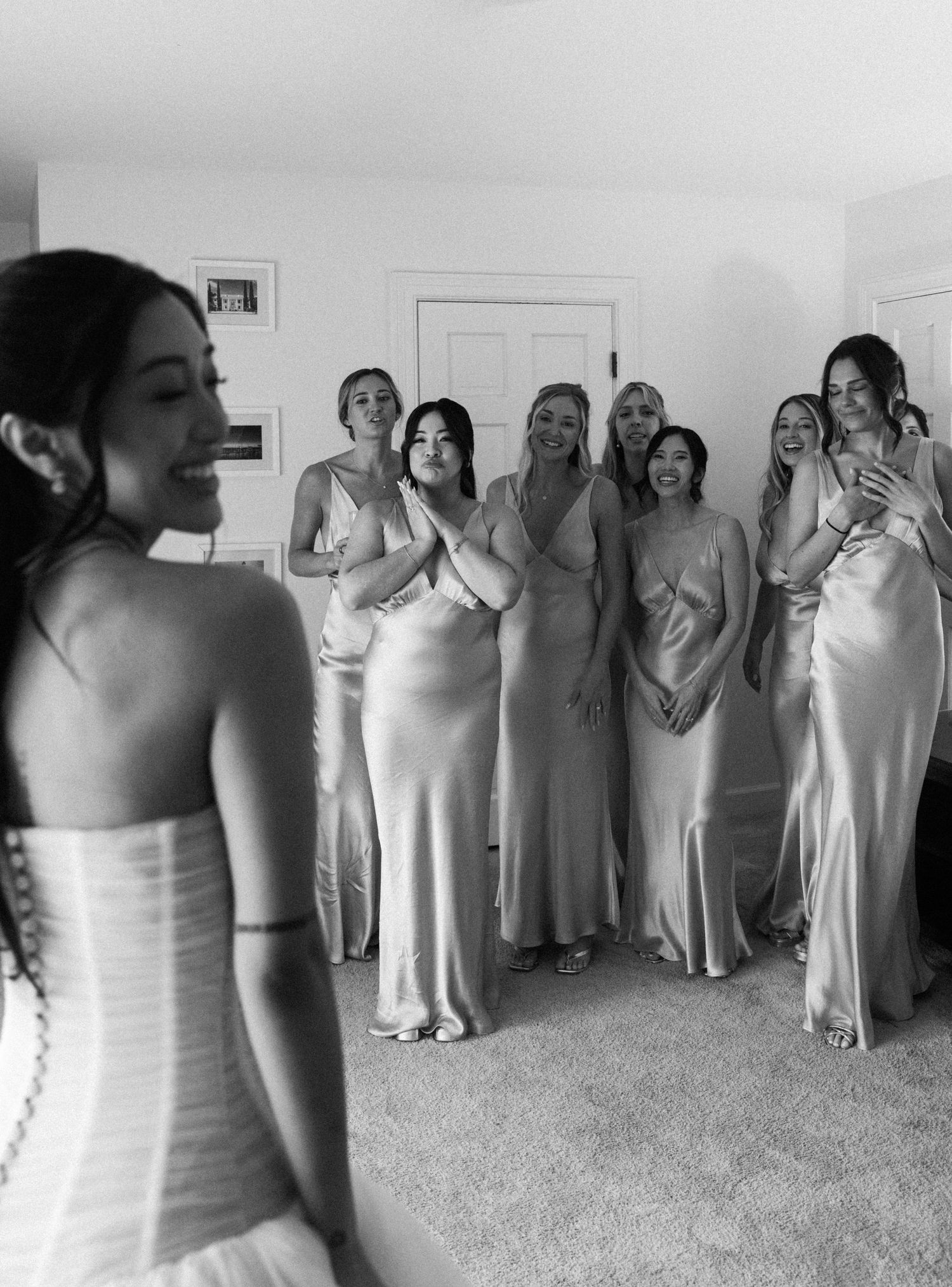 Black and white photo of bridesmaids in satin gowns reacting emotionally to bride during wedding morning reveal.