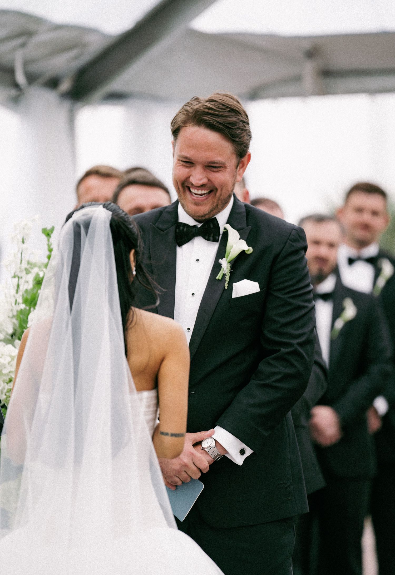 Joyful groom in black tuxedo laughs while holding hands with bride during outdoor wedding ceremony.