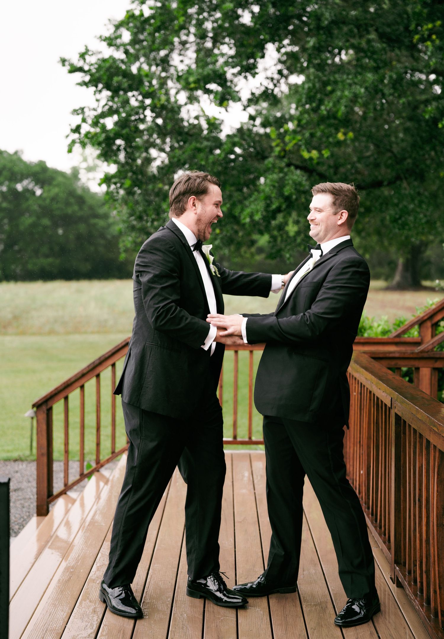 Two groomsmen in black tuxedos laughing and shaking hands on wooden deck outdoors.