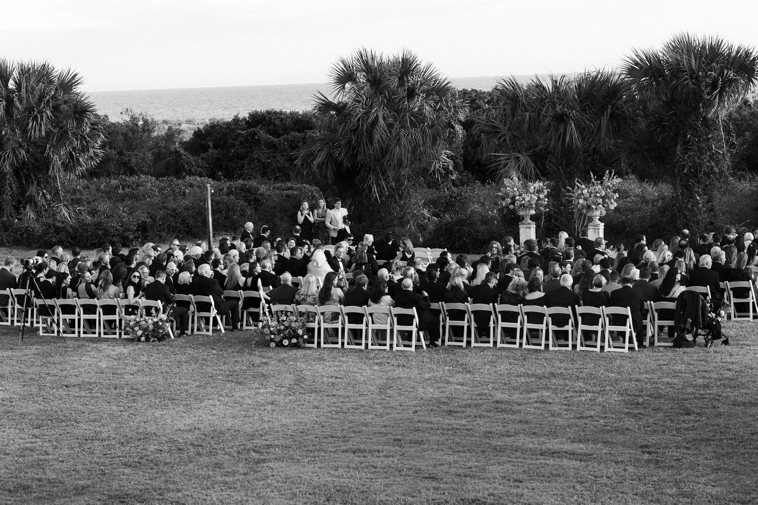 Black and white aerial view of outdoor wedding reception with guests seated at long tables on green lawn.
