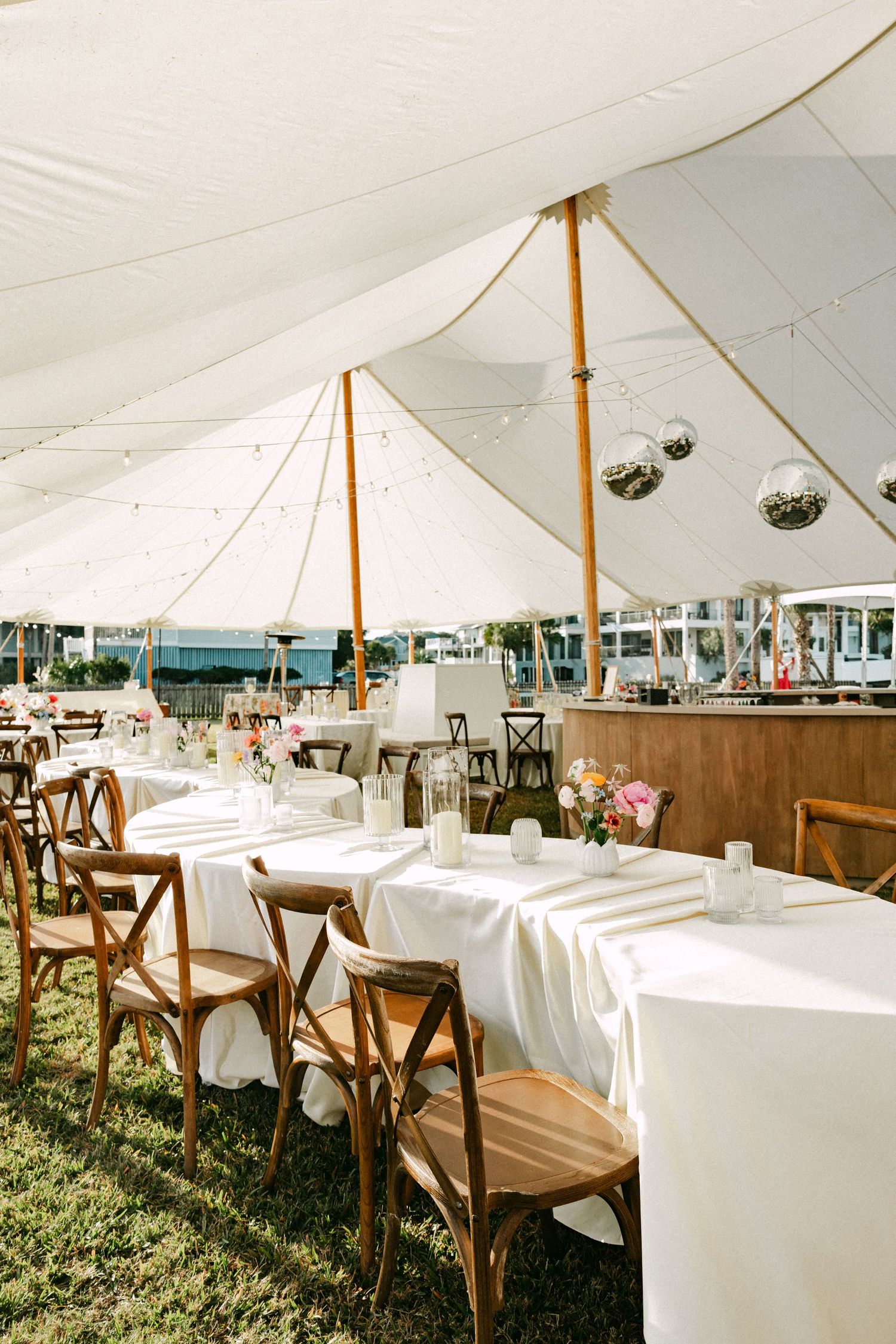 Sunlit sailcloth wedding reception tent with wooden crossback chairs, white linen tables, and hanging disco balls.