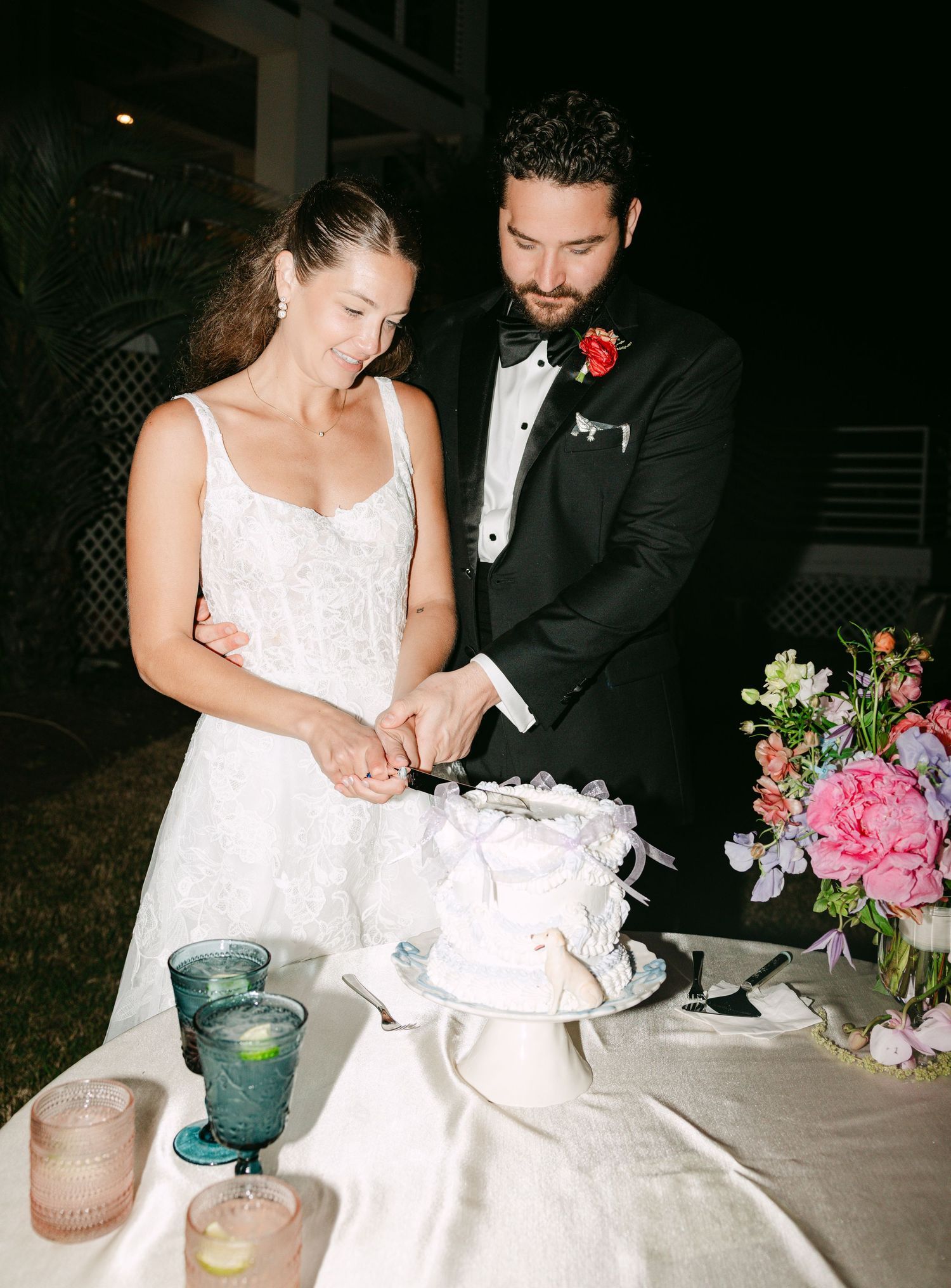 Bride and groom cut white wedding cake together at candlelit outdoor reception table at night.