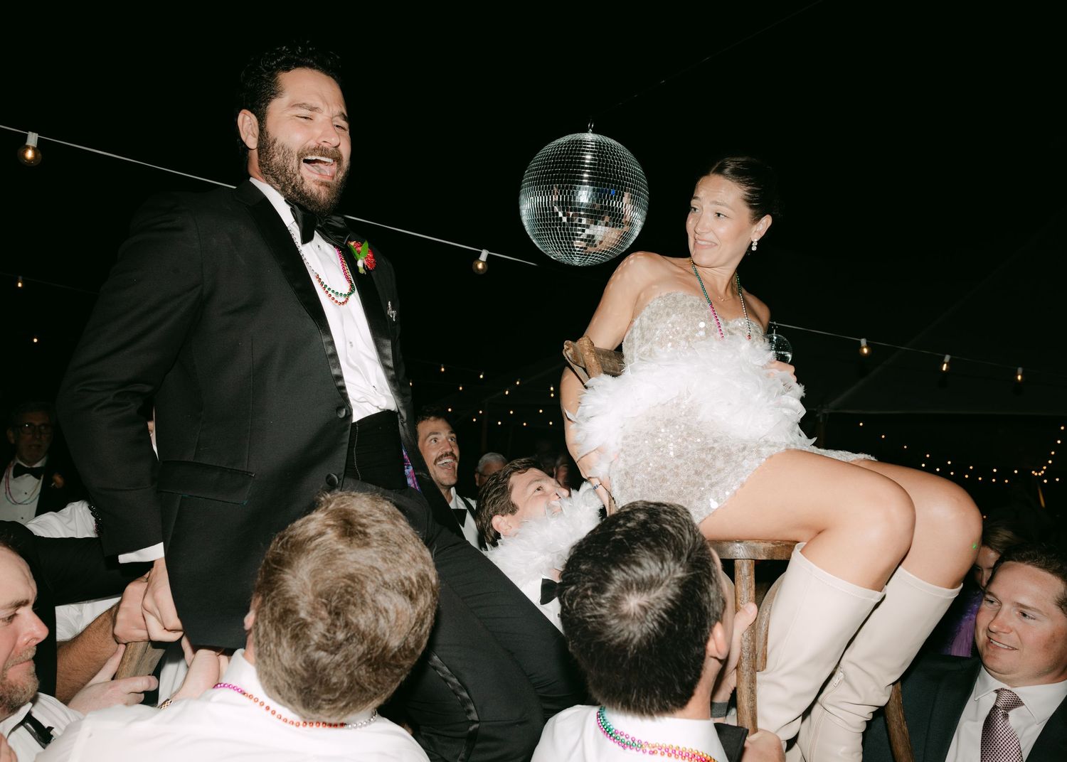Bride in white feathered dress lifted by guests during lively wedding reception celebration under disco ball.
