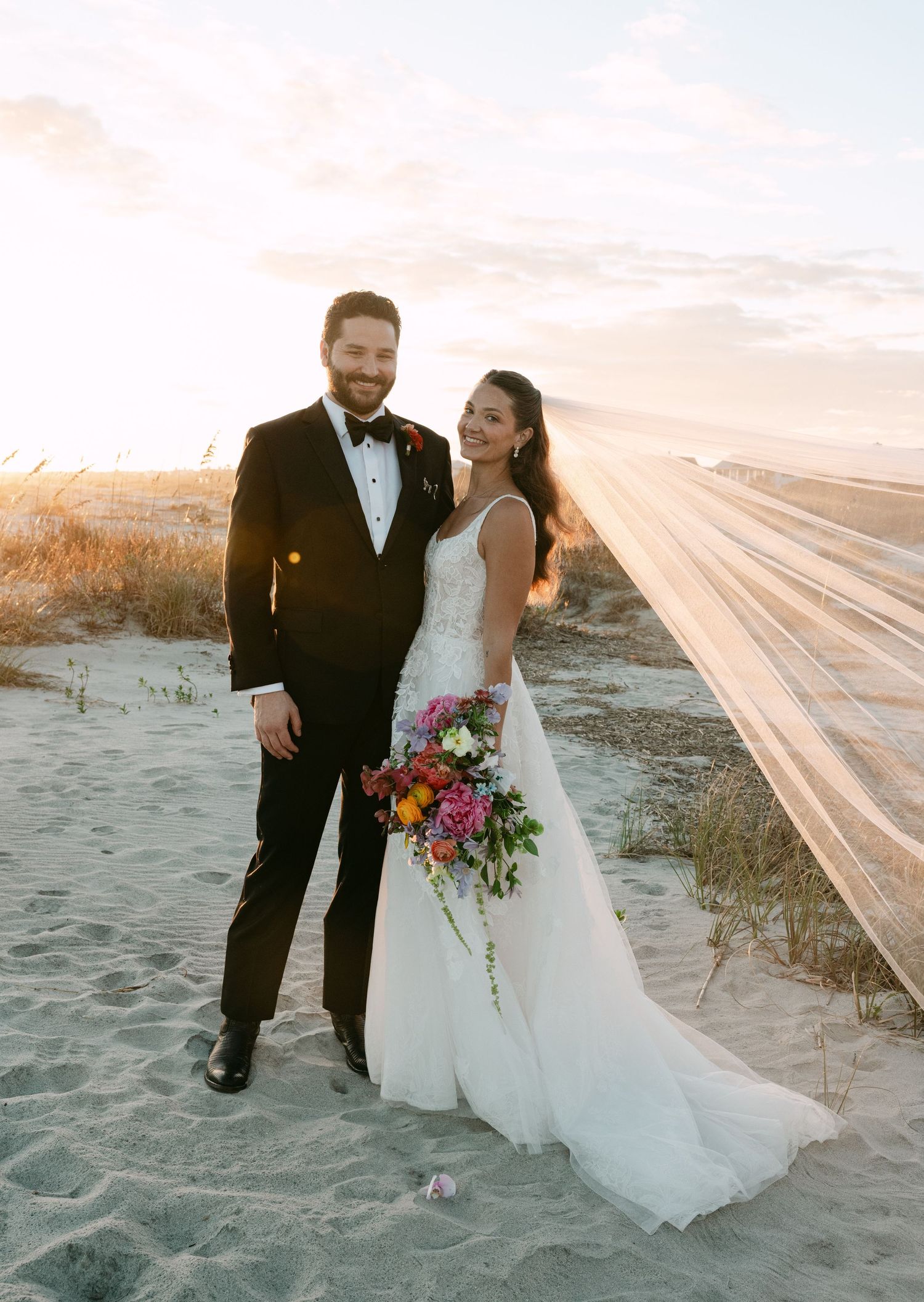 Bride and groom pose on sandy beach at sunset, her veil billowing in the wind, holding colorful bridal bouquet.
