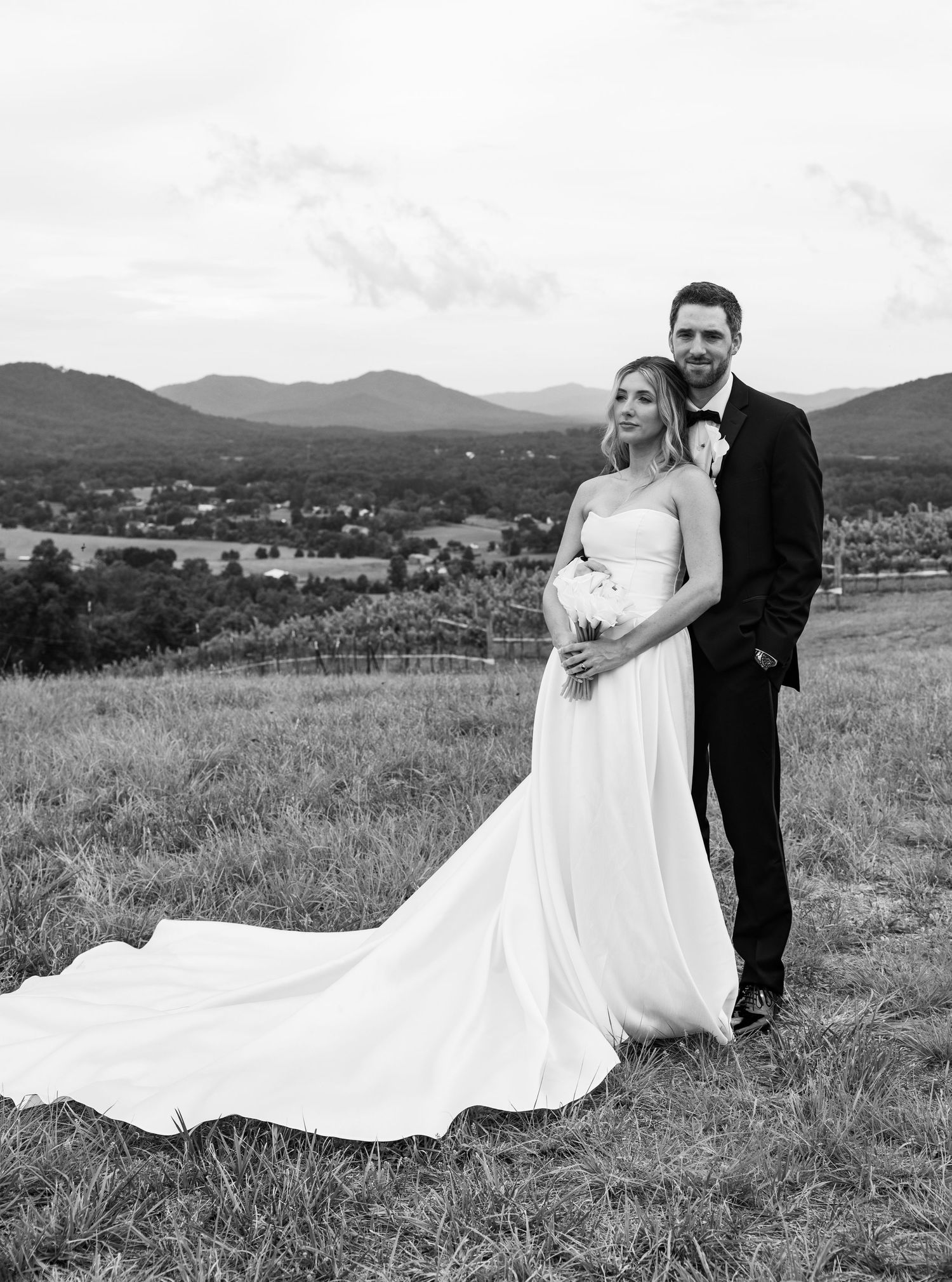 Black and white portrait of bride and groom embracing on hilltop with stunning mountain valley backdrop.
