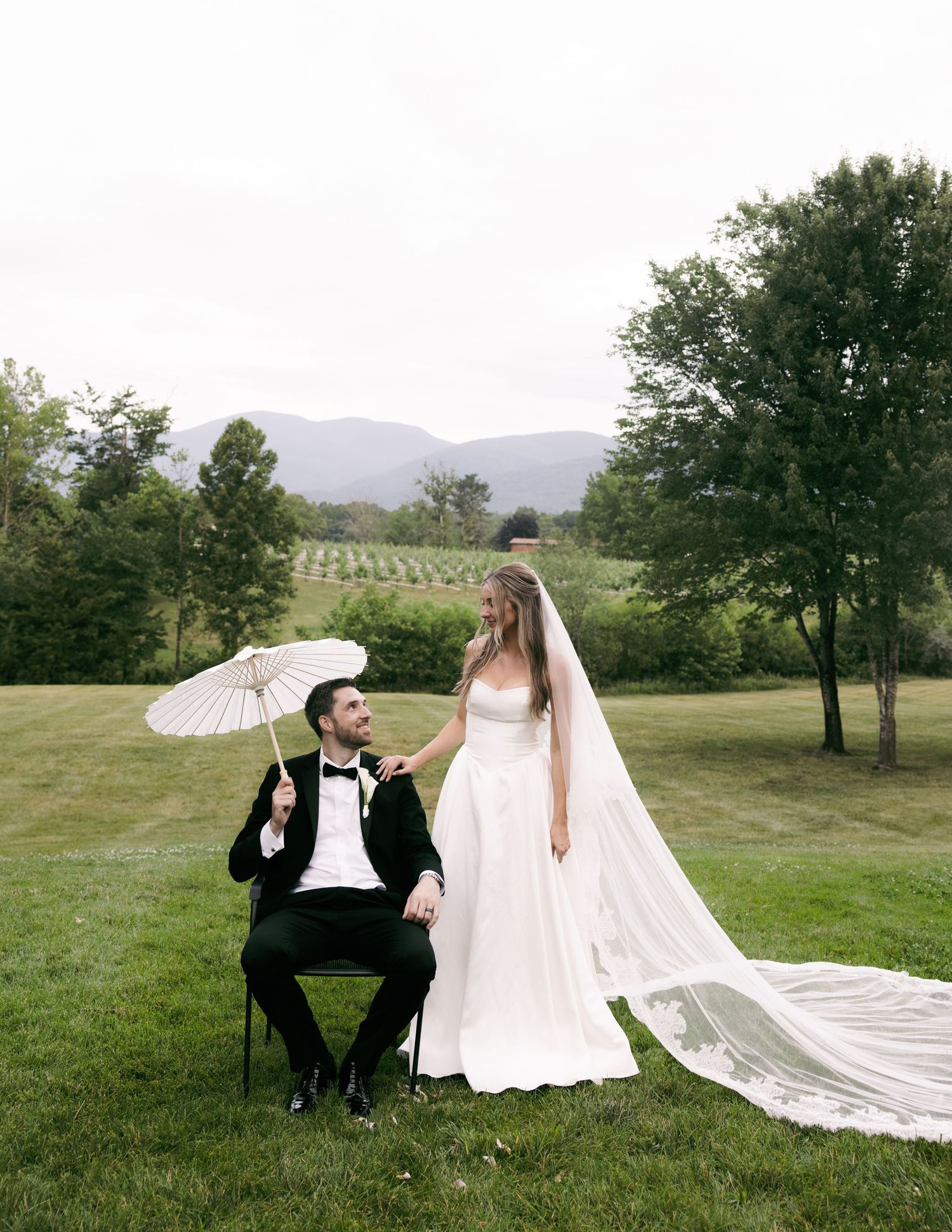Bride and groom pose on lush green lawn with mountain vineyard views, groom holding white parasol.