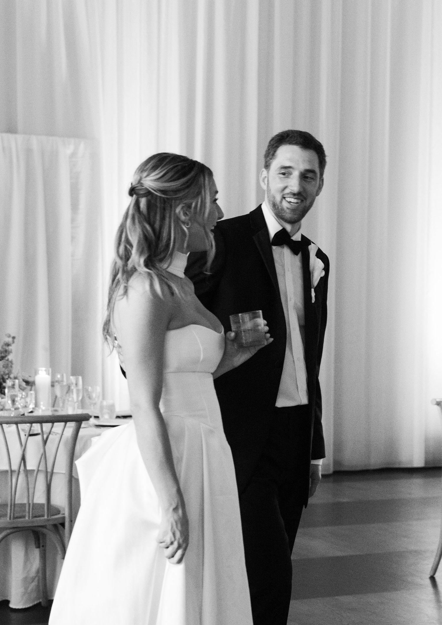 Black and white photo of bride and groom smiling together at elegant white-draped wedding reception.