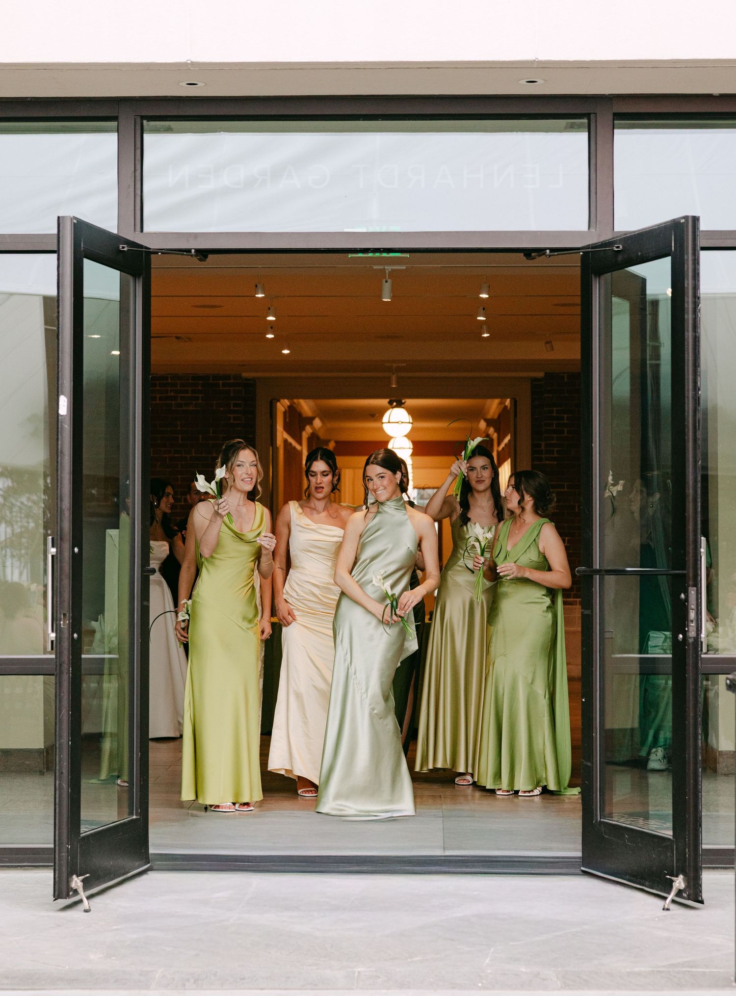Bridesmaids in green satin dresses stand with bride in doorway of modern wedding venue.