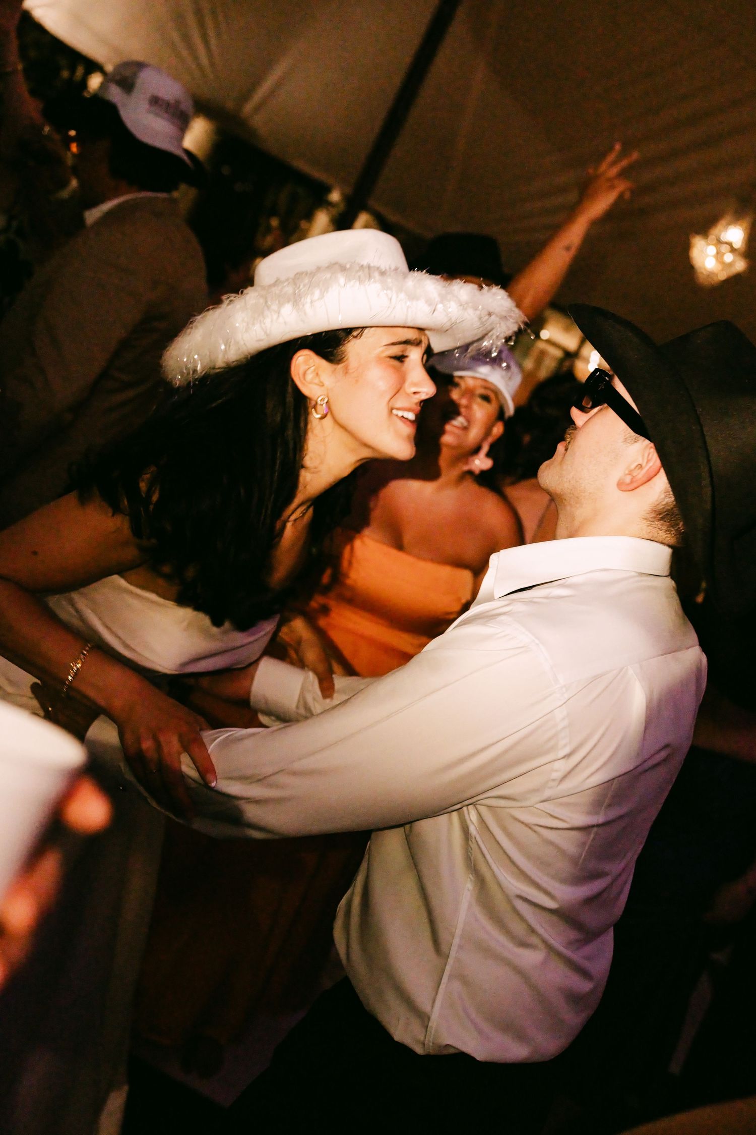 Bride in white feathered hat dancing closely with groom in white shirt at wedding reception.