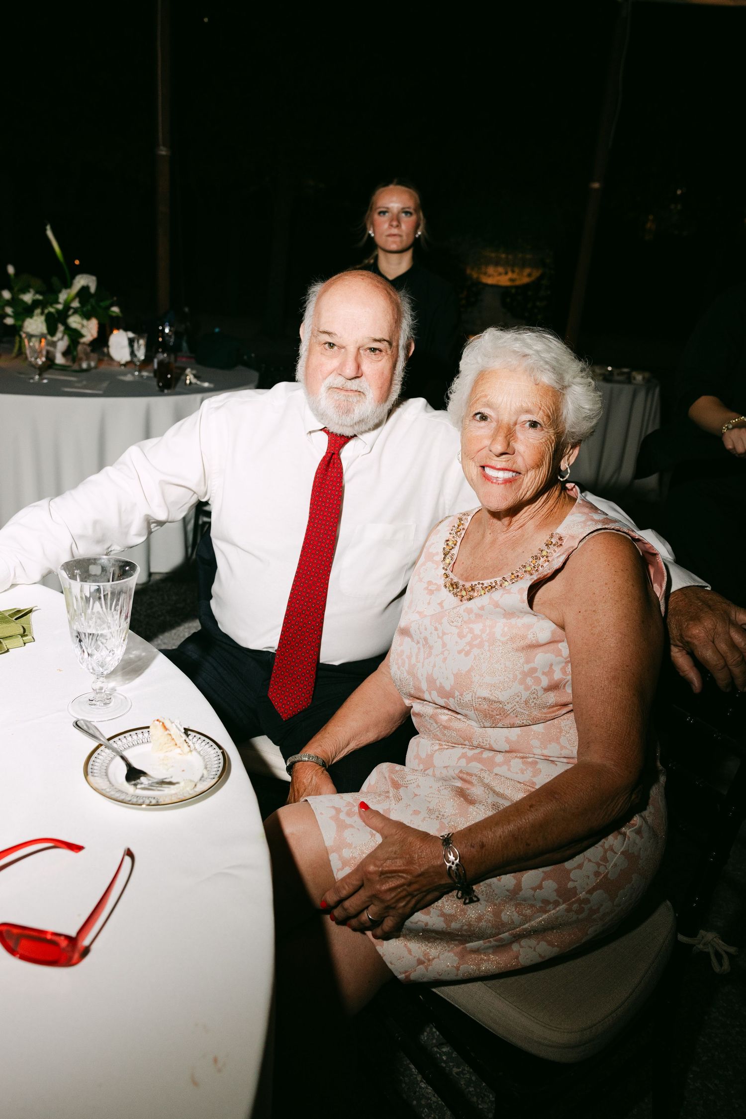 Older couple smiles at wedding reception table with candles and red-framed glasses nearby.