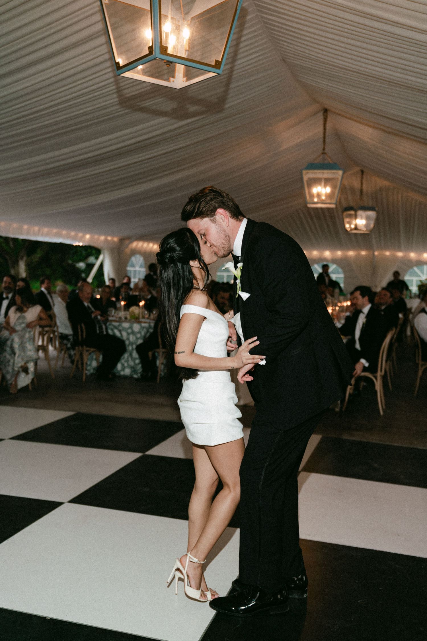 Bride and groom share a romantic kiss on a black and white checkered dance floor under a elegant tented wedding reception.