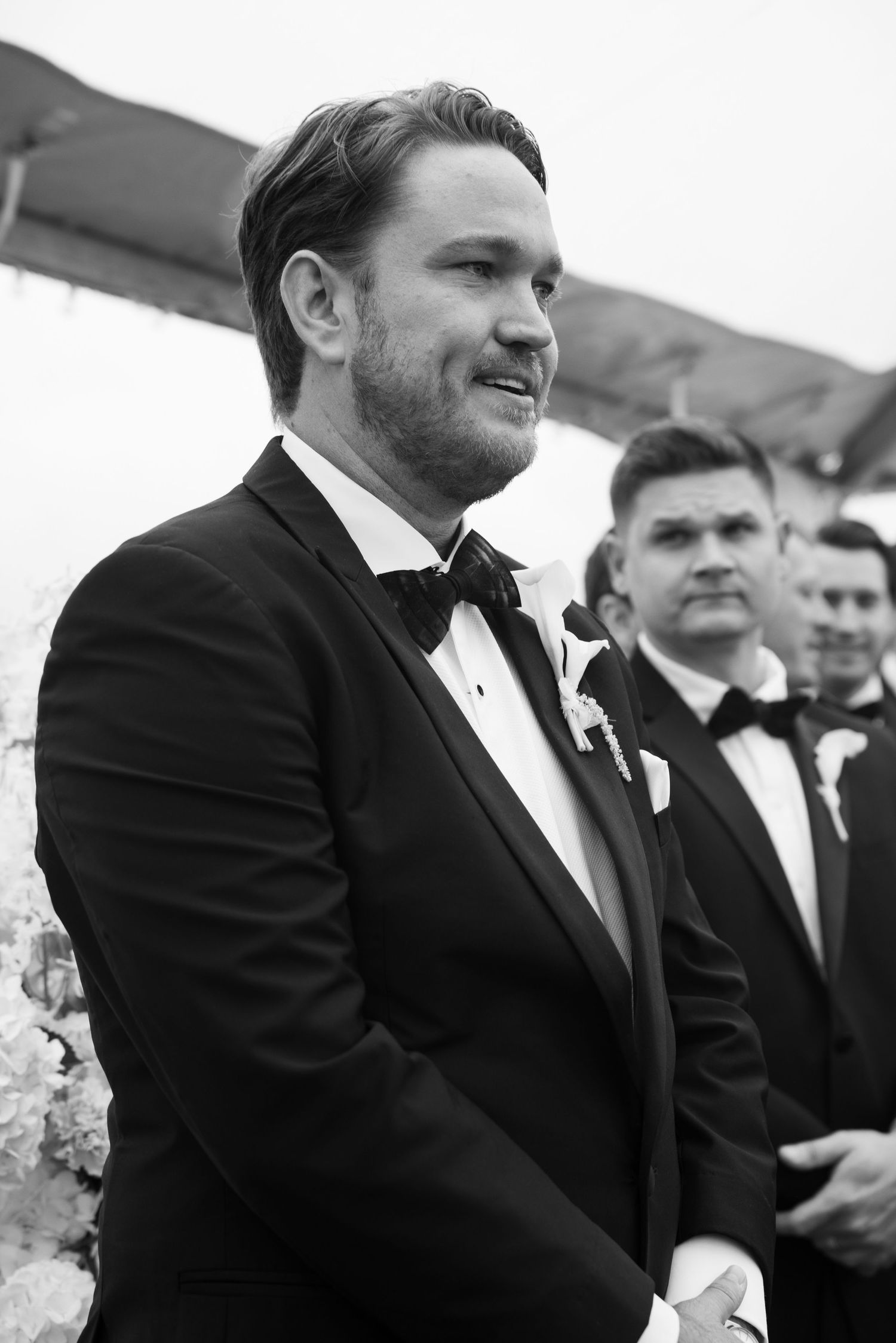 Groom in black tuxedo and bow tie smiling at wedding ceremony, black and white photo.
