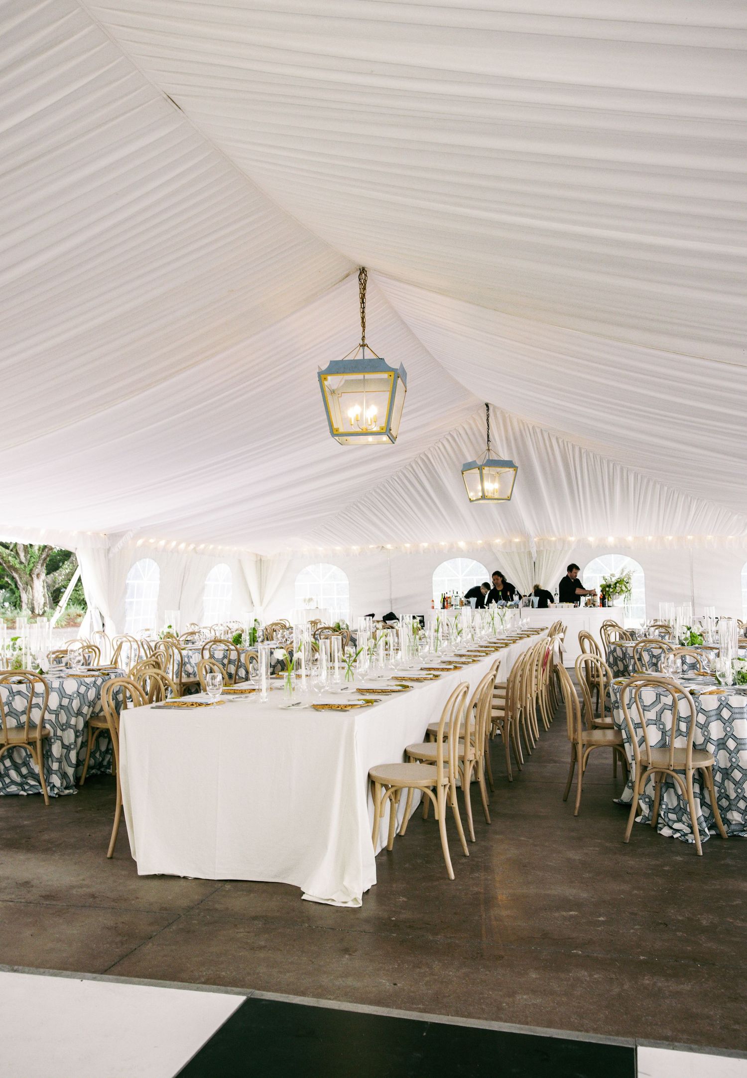 Elegant tented wedding reception with long white tables, gold chairs, and hanging lanterns.