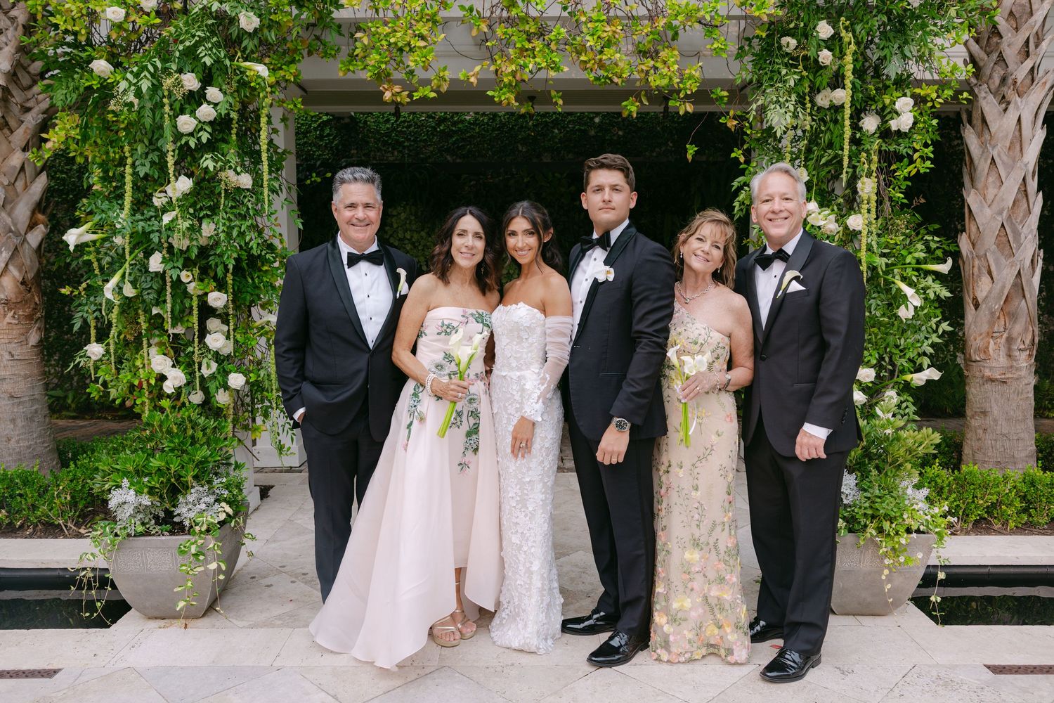 Wedding party gathered for formal photos under palm trees and white columns.