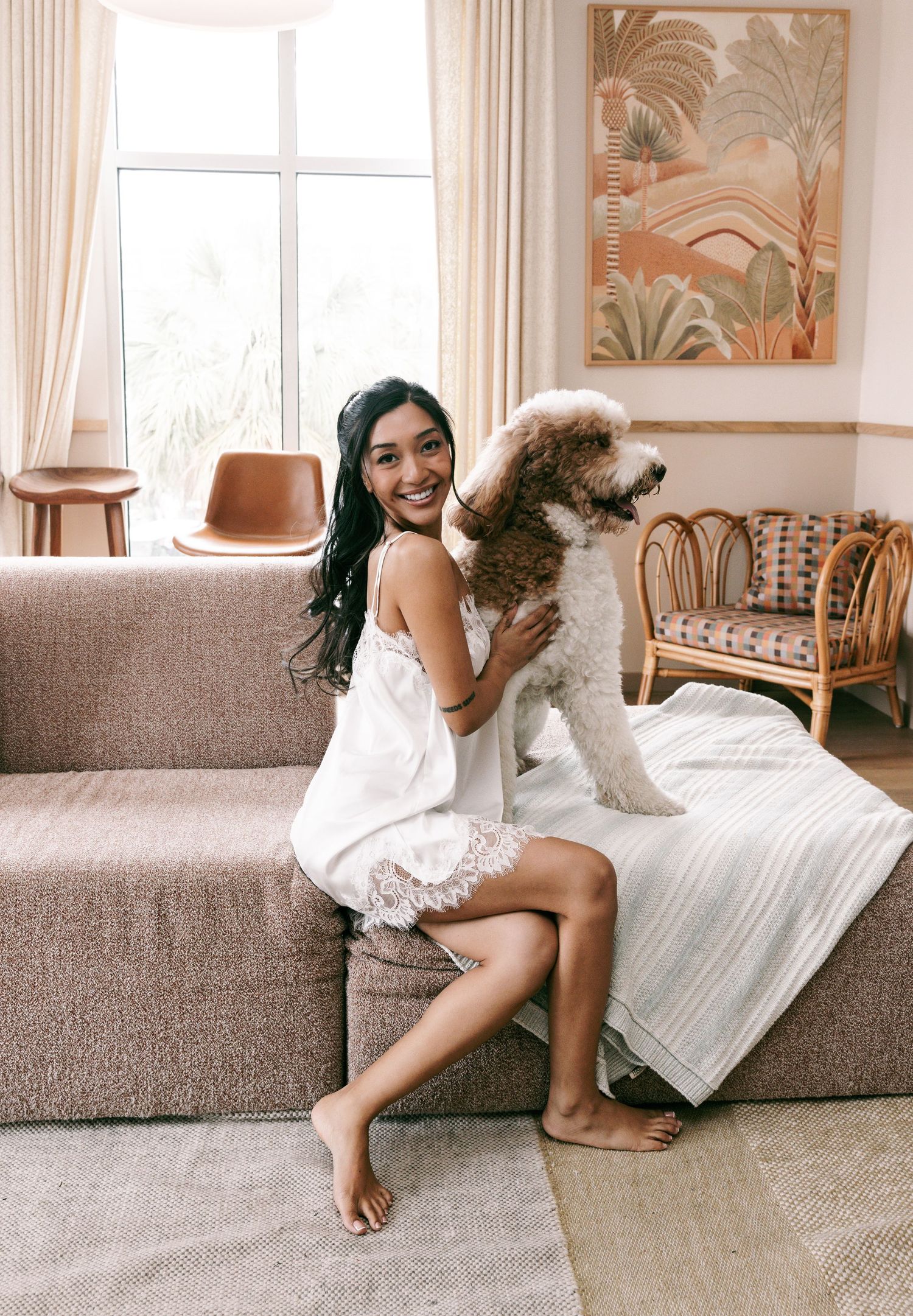 Young woman in white dress smiling on pink sofa with fluffy brown dog in cozy living room.