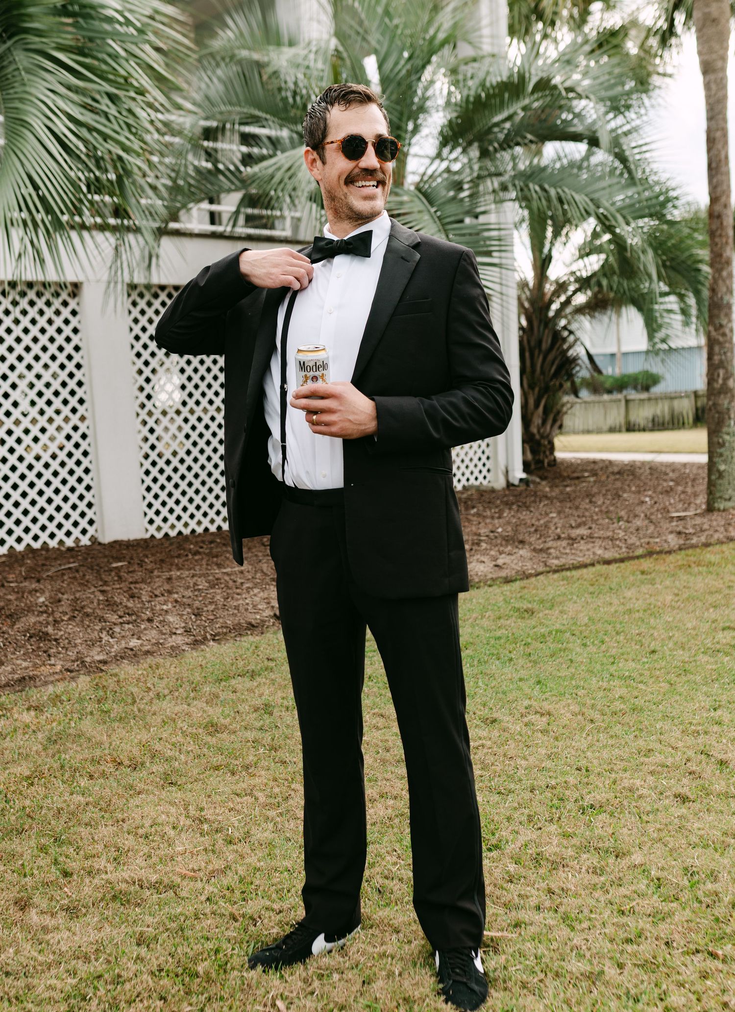 Smiling groomsman in black tuxedo with sunglasses holding beer can outdoors.