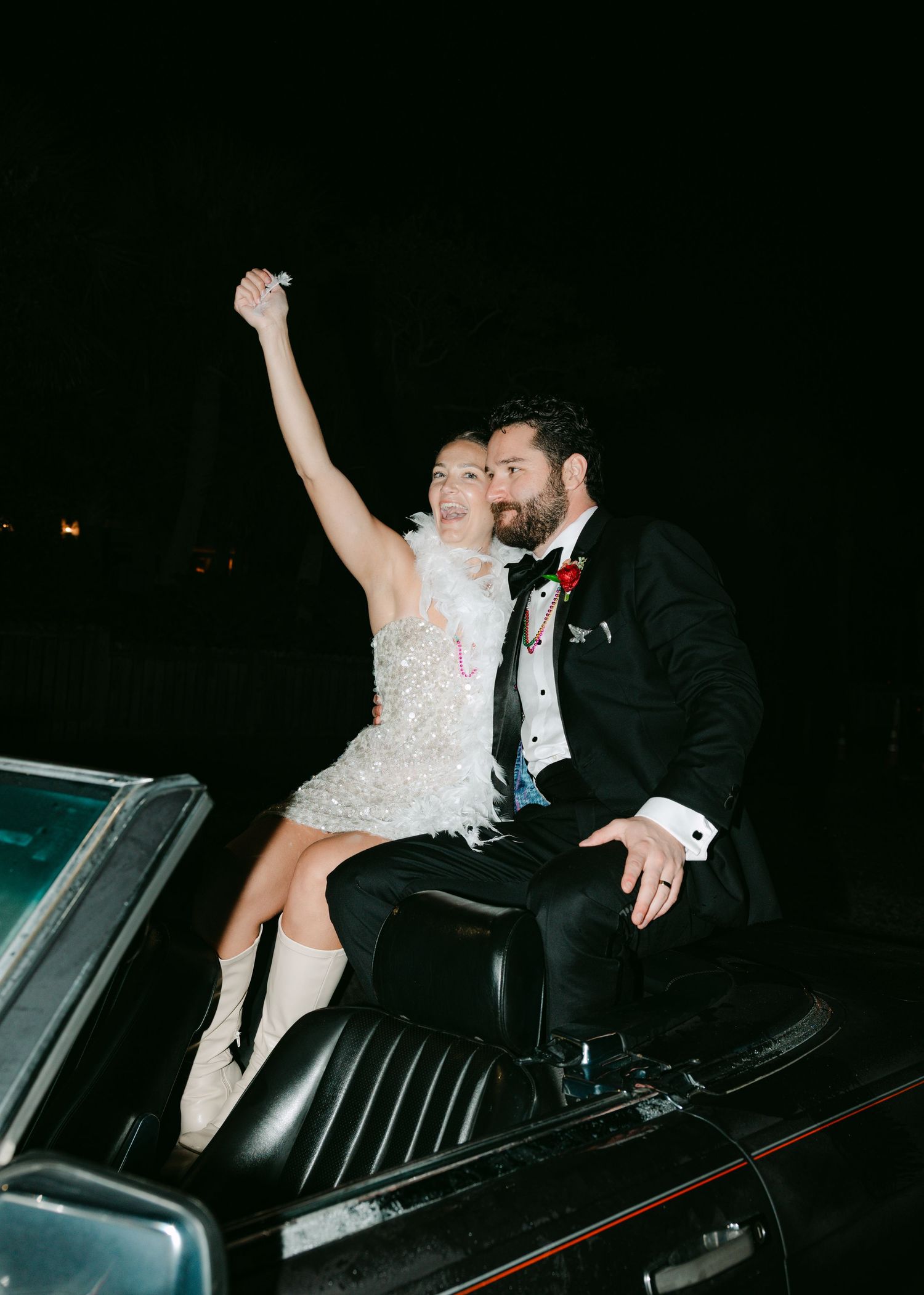 Bride in sparkly mini dress raises fist while groom kisses her cheek seated on vintage convertible.