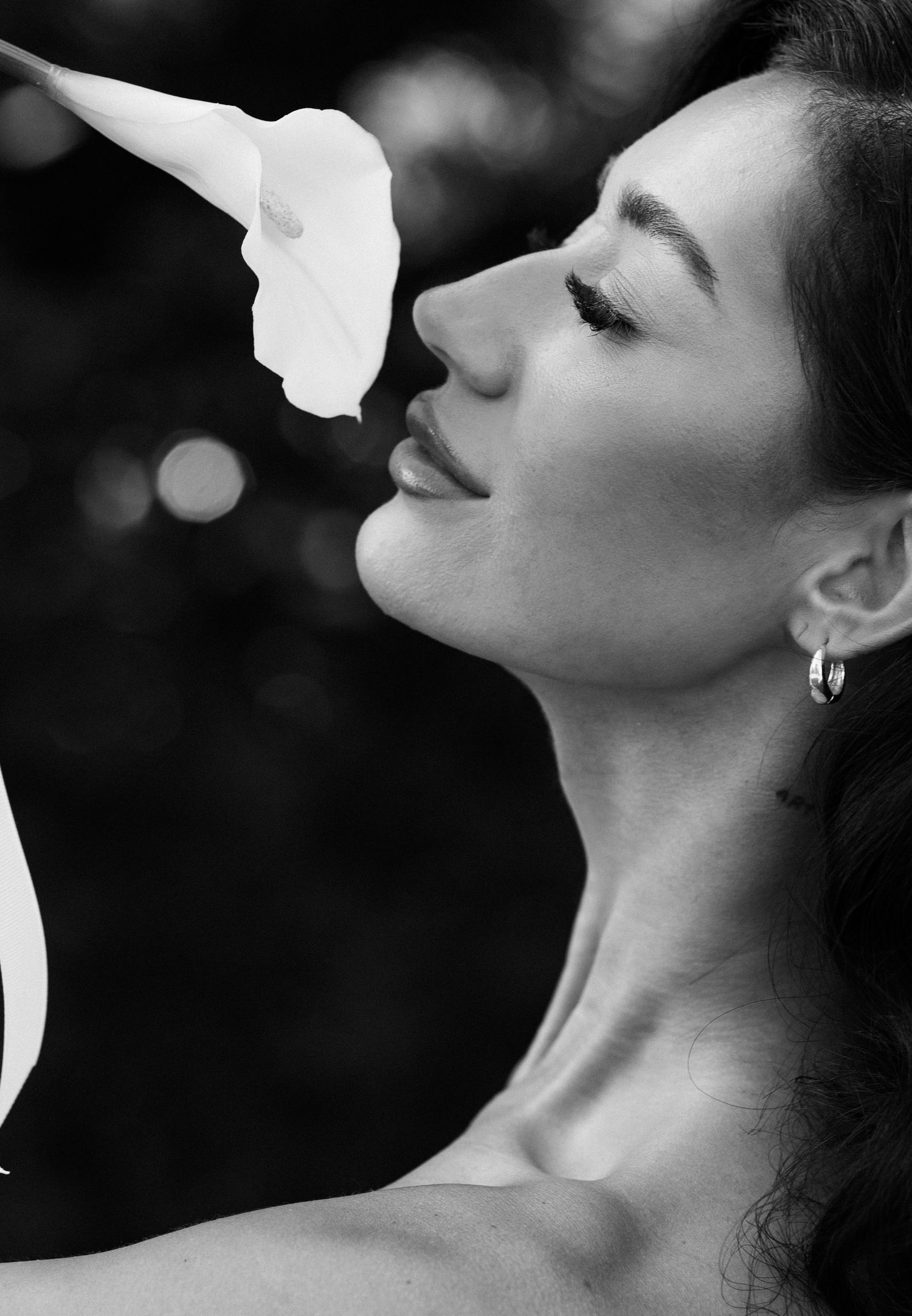 Black and white profile portrait of a woman smelling a white calla lily flower, wearing hoop earrings.