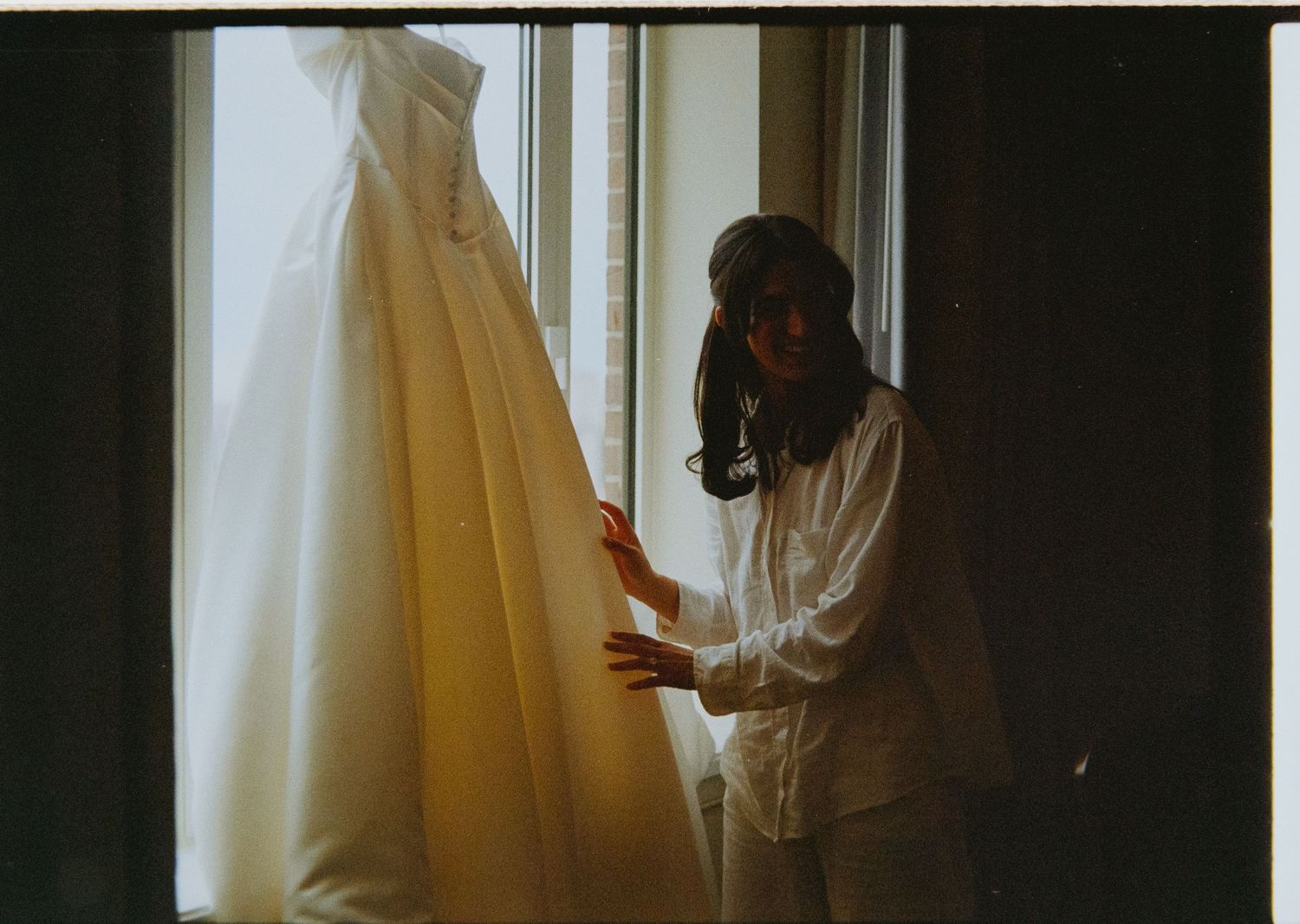 Bride admiring her wedding gown hanging by a sunlit window before the ceremony.