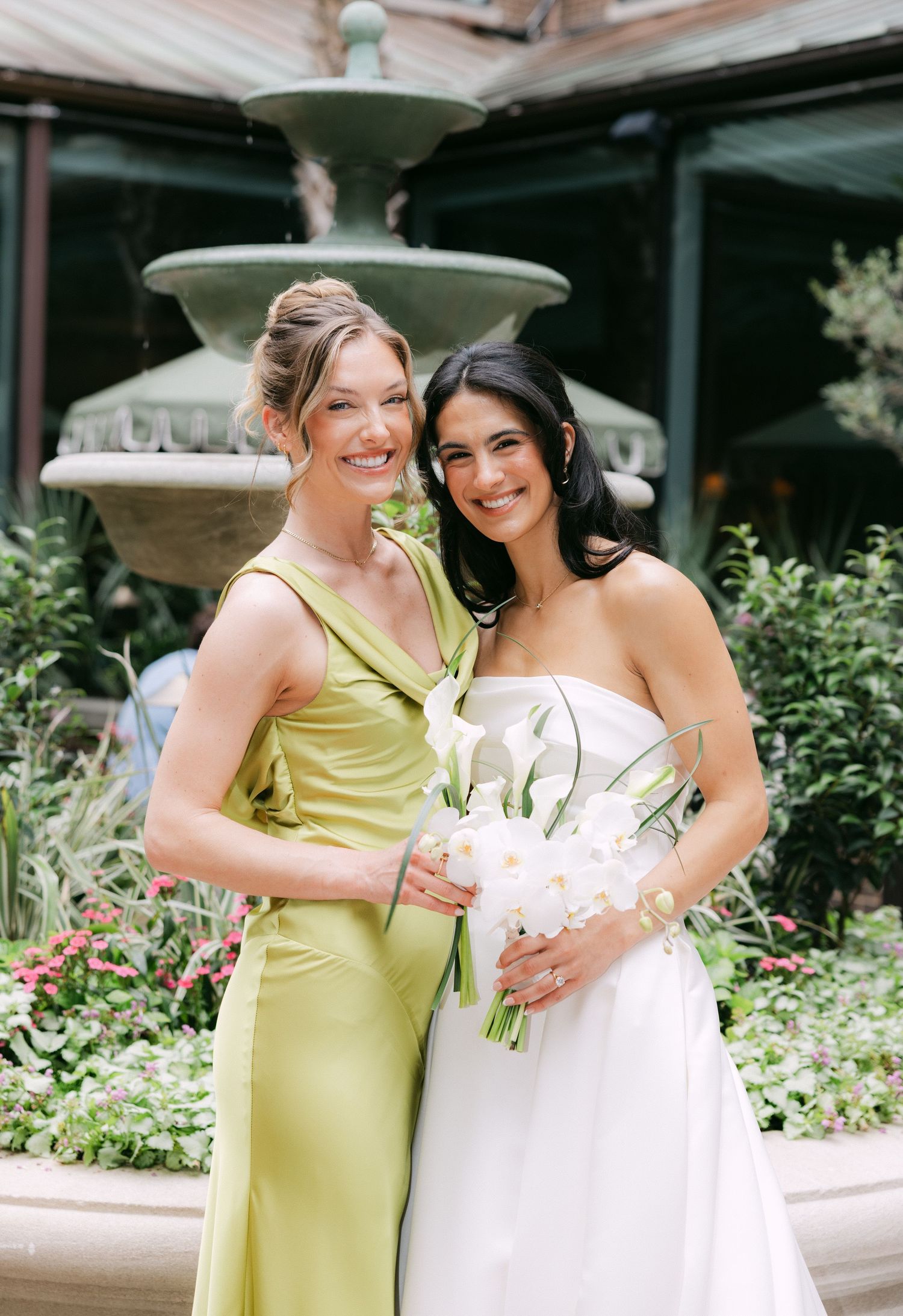 Bride in white strapless gown holds calla lily bouquet beside bridesmaid in chartreuse dress near garden fountain.