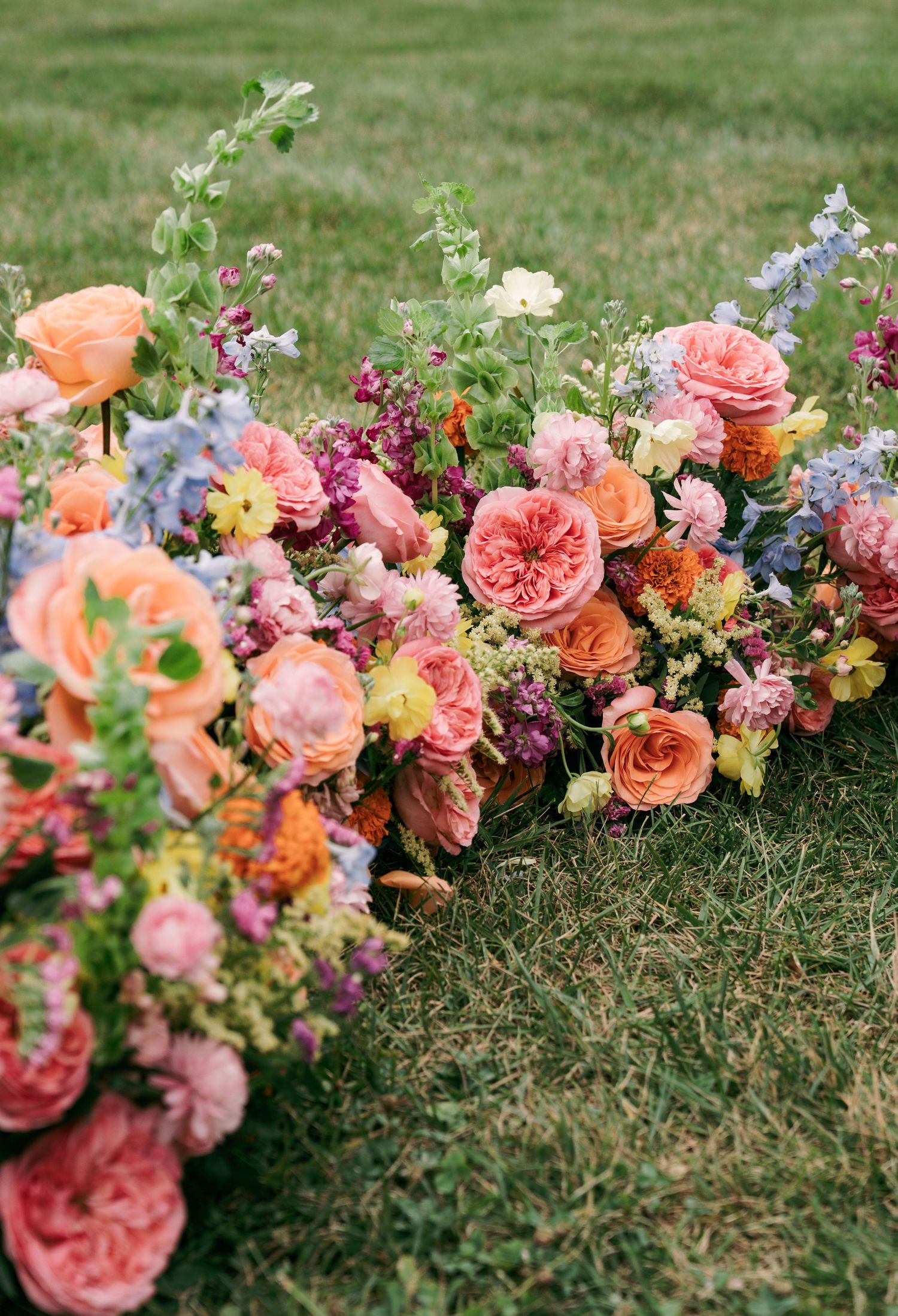Lush garden floral arrangements with peach roses, pink peonies, and blue delphiniums on green grass.