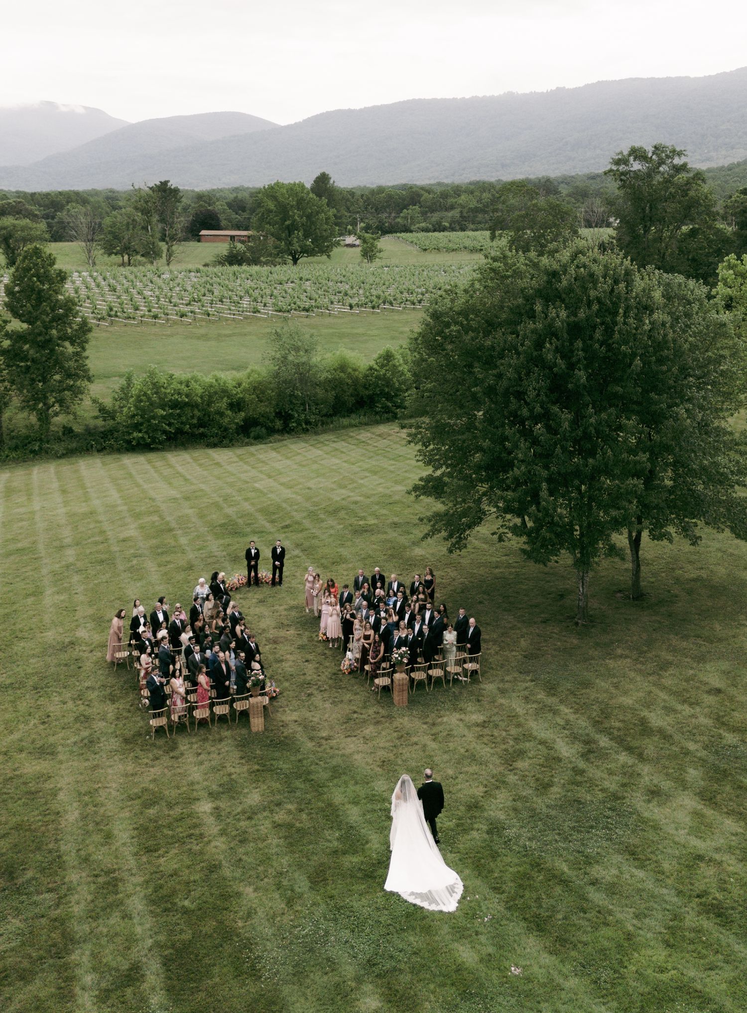 Aerial view of an outdoor wedding ceremony on a lush green lawn with mountain backdrop and guests seated in rows.