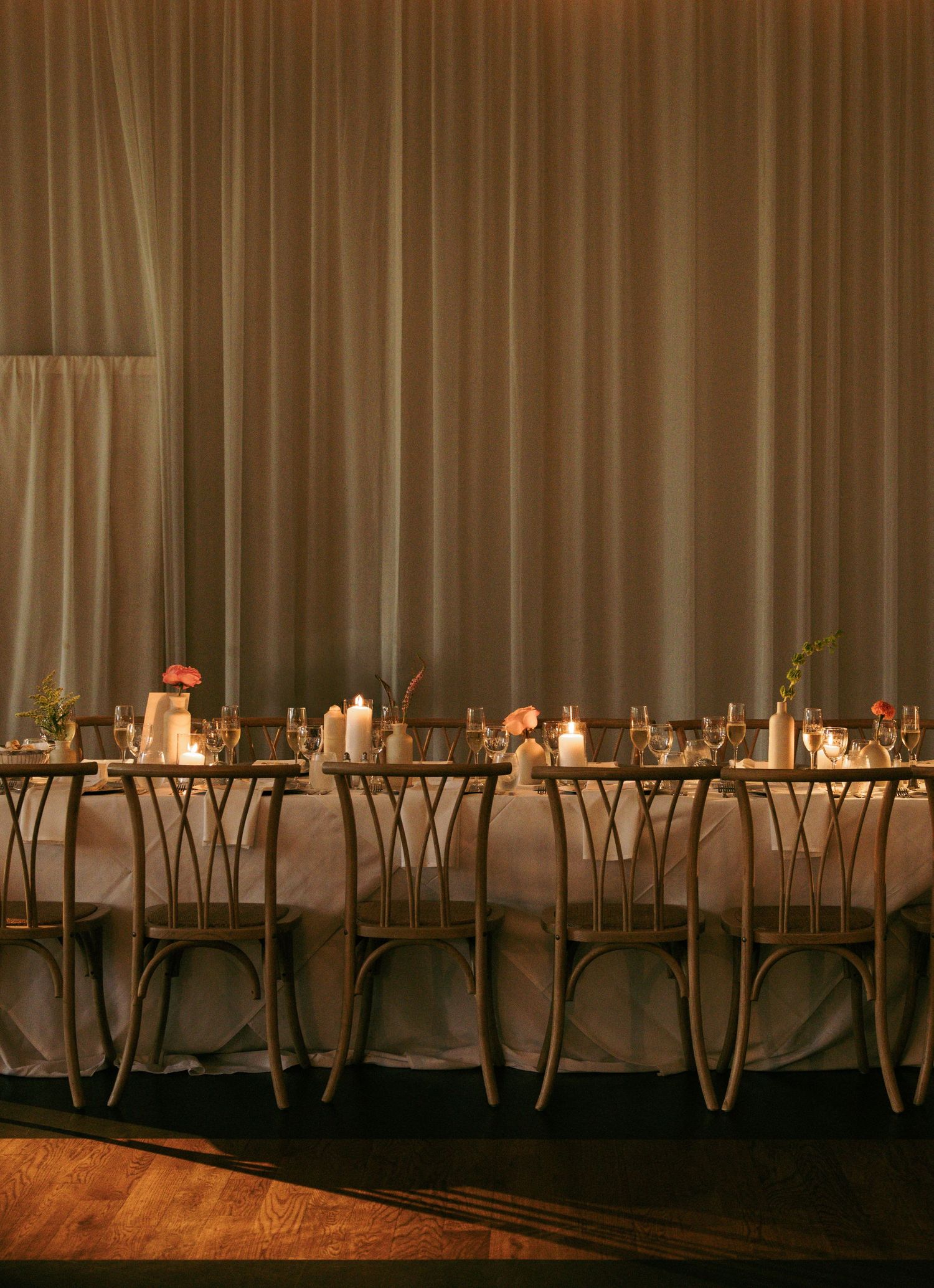 Elegant candlelit reception table with gold chairs, floral centerpieces, and draped fabric backdrop.