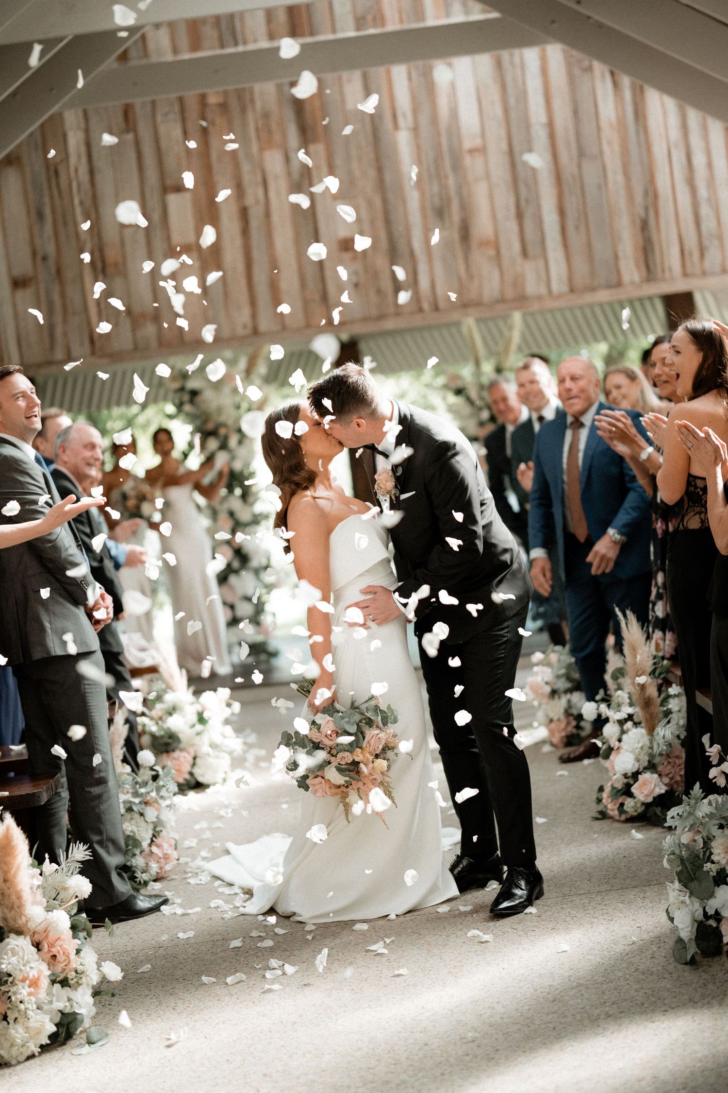 A tender wedding moment captured with white rose petals falling as guests watch at an elegant indoor ceremony space.