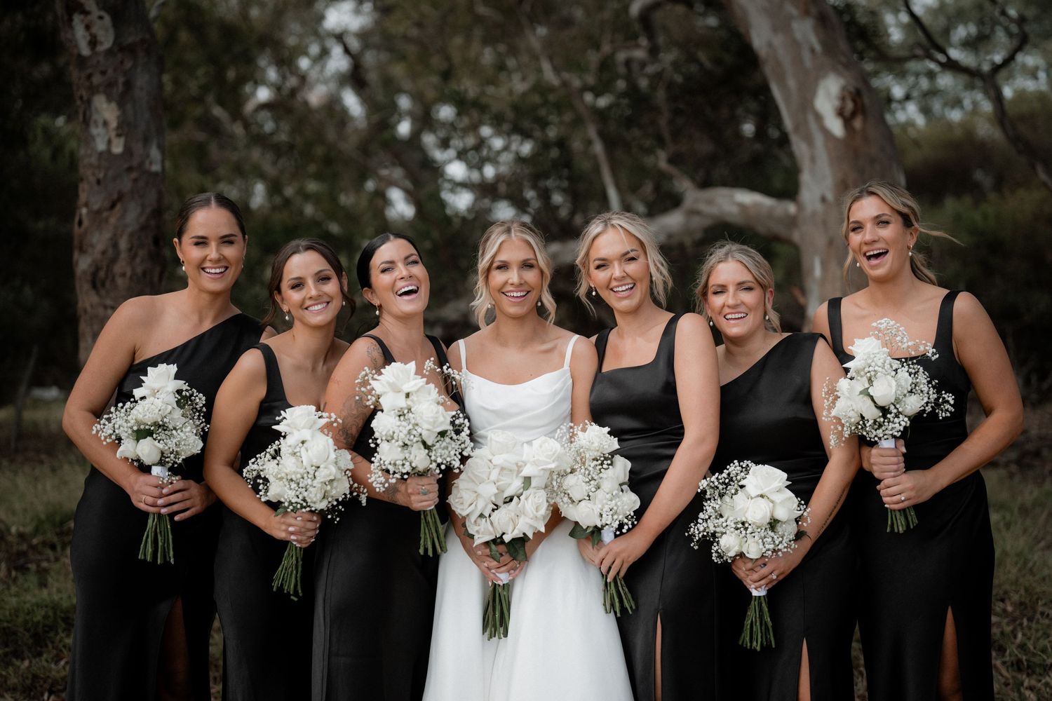 Wedding party in elegant black bridesmaid dresses holding white bouquets pose together outdoors under trees.