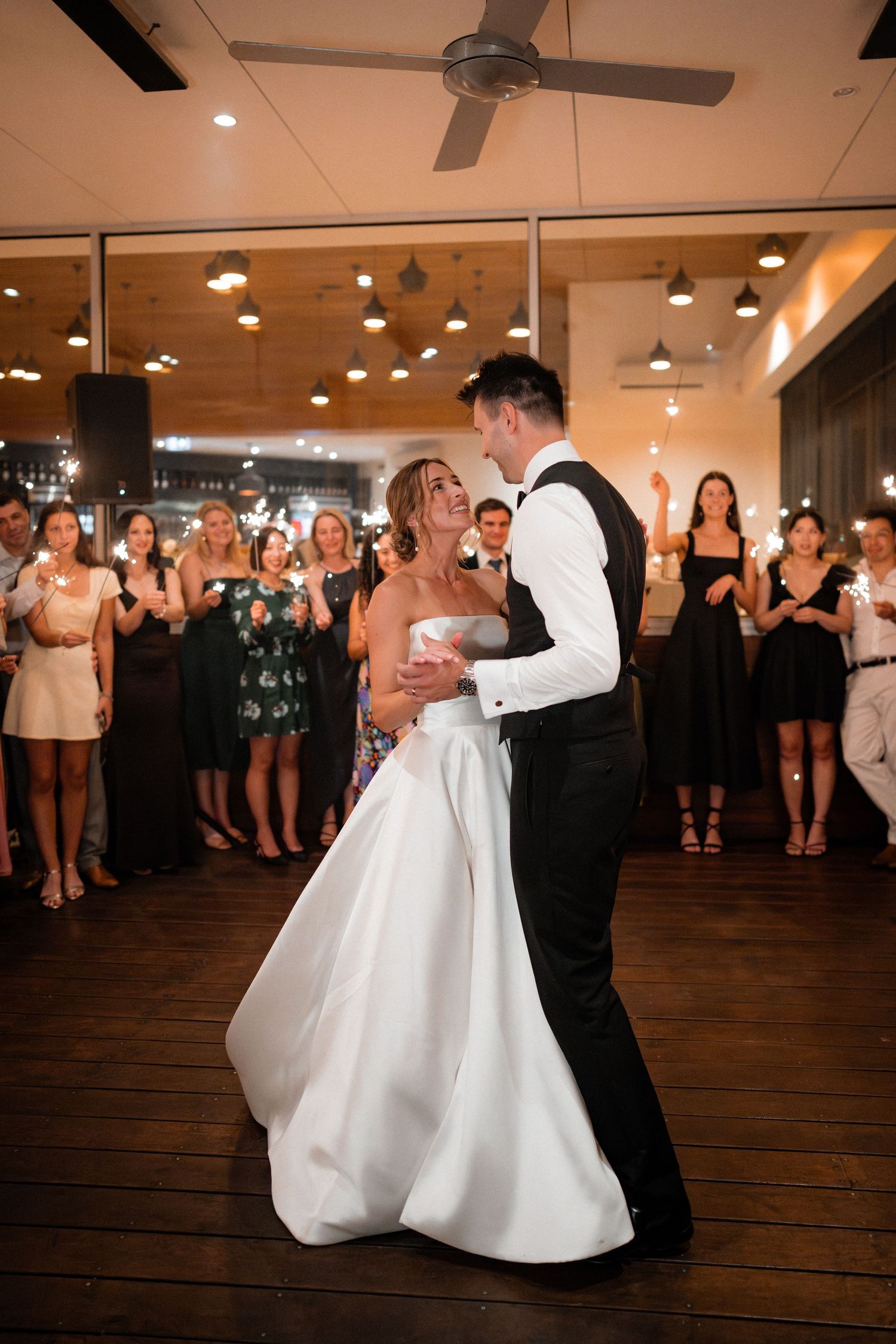A couple shares their first dance surrounded by wedding guests in an indoor reception venue with warm lighting.