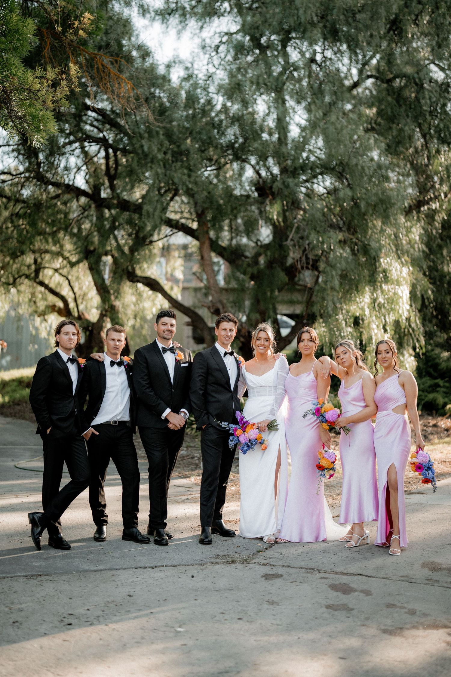 Wedding party in black and lavender pose playfully together in an outdoor setting beneath trees.