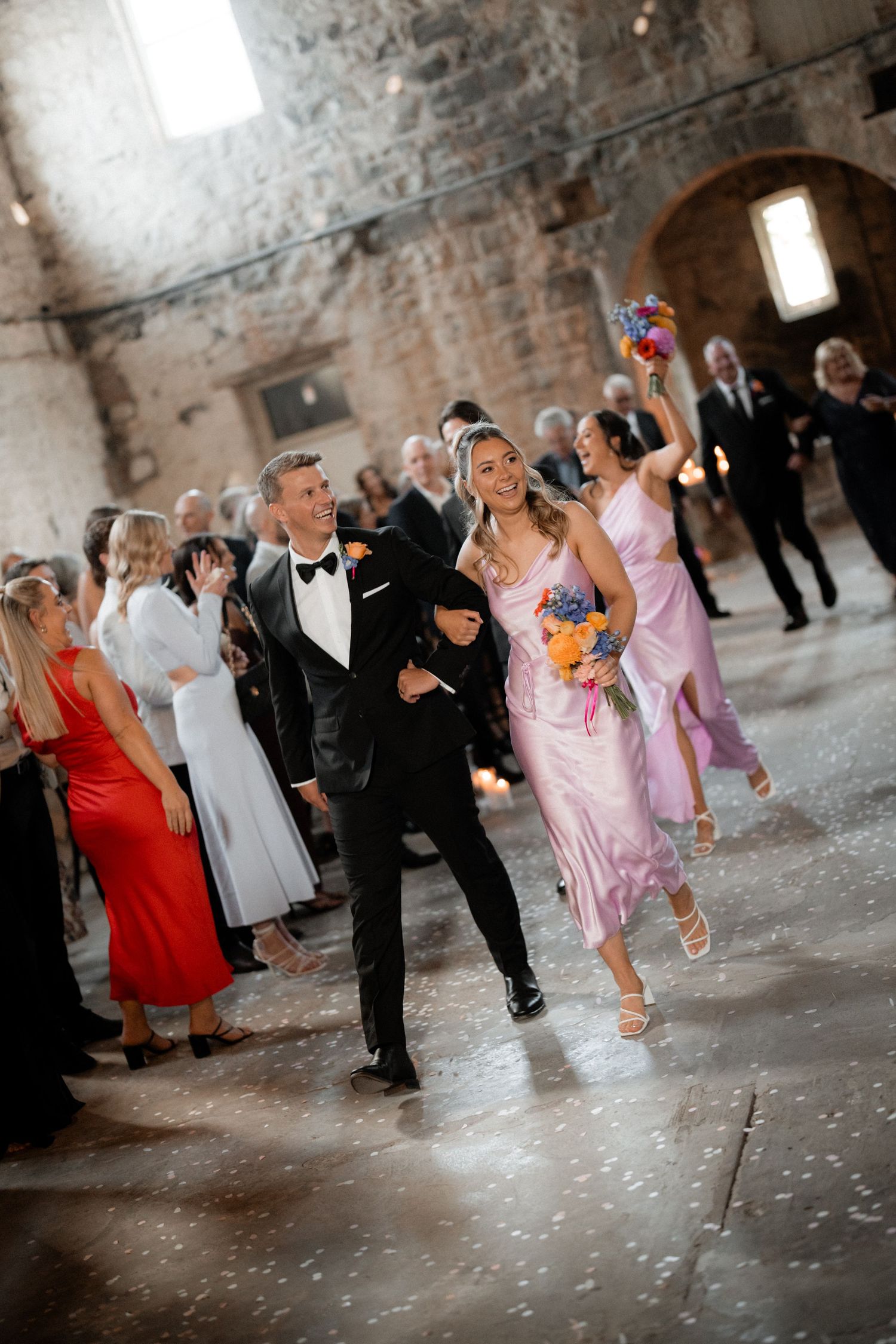 Wedding celebration in a rustic stone venue with guests in formal attire and colorful bouquets.