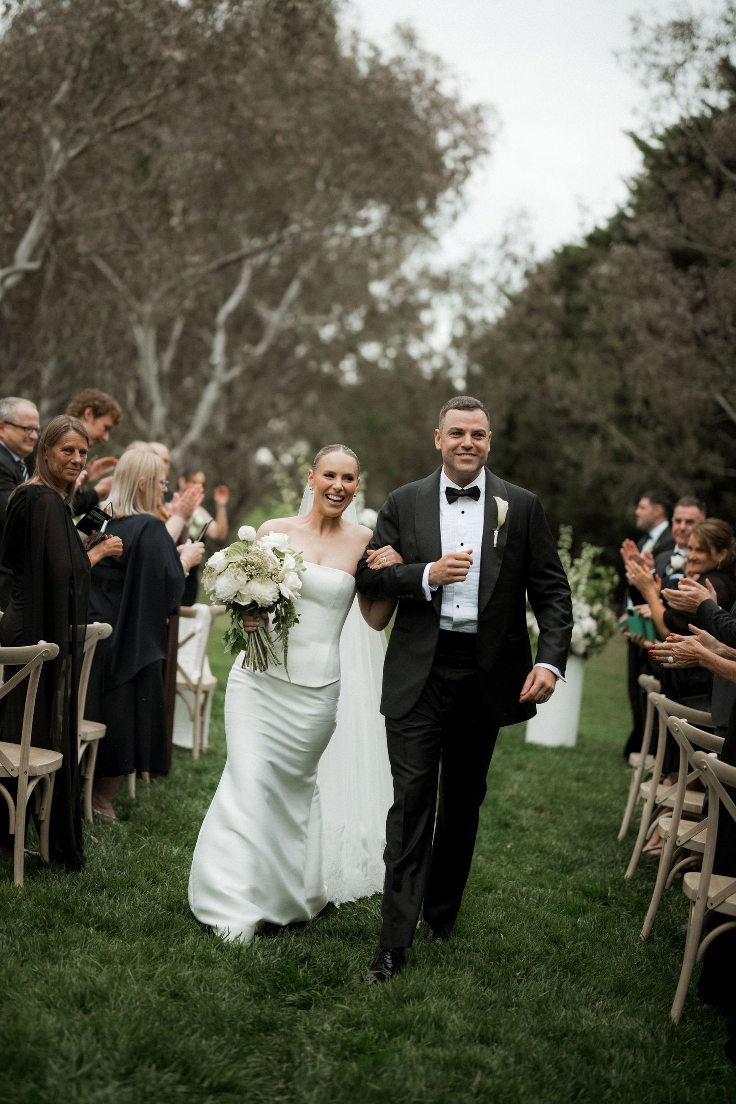 Bride and groom walk joyfully down outdoor wedding aisle lined with guests, bride holding white bouquet.