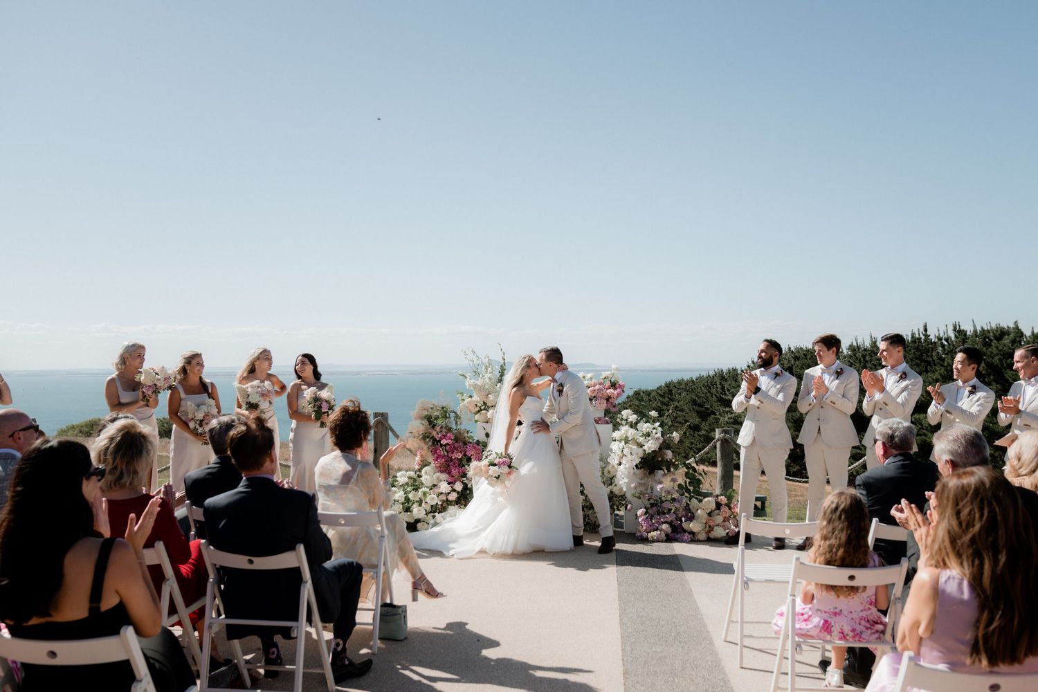 Wedding guests gather outdoors to celebrate during a sunny coastal ceremony.