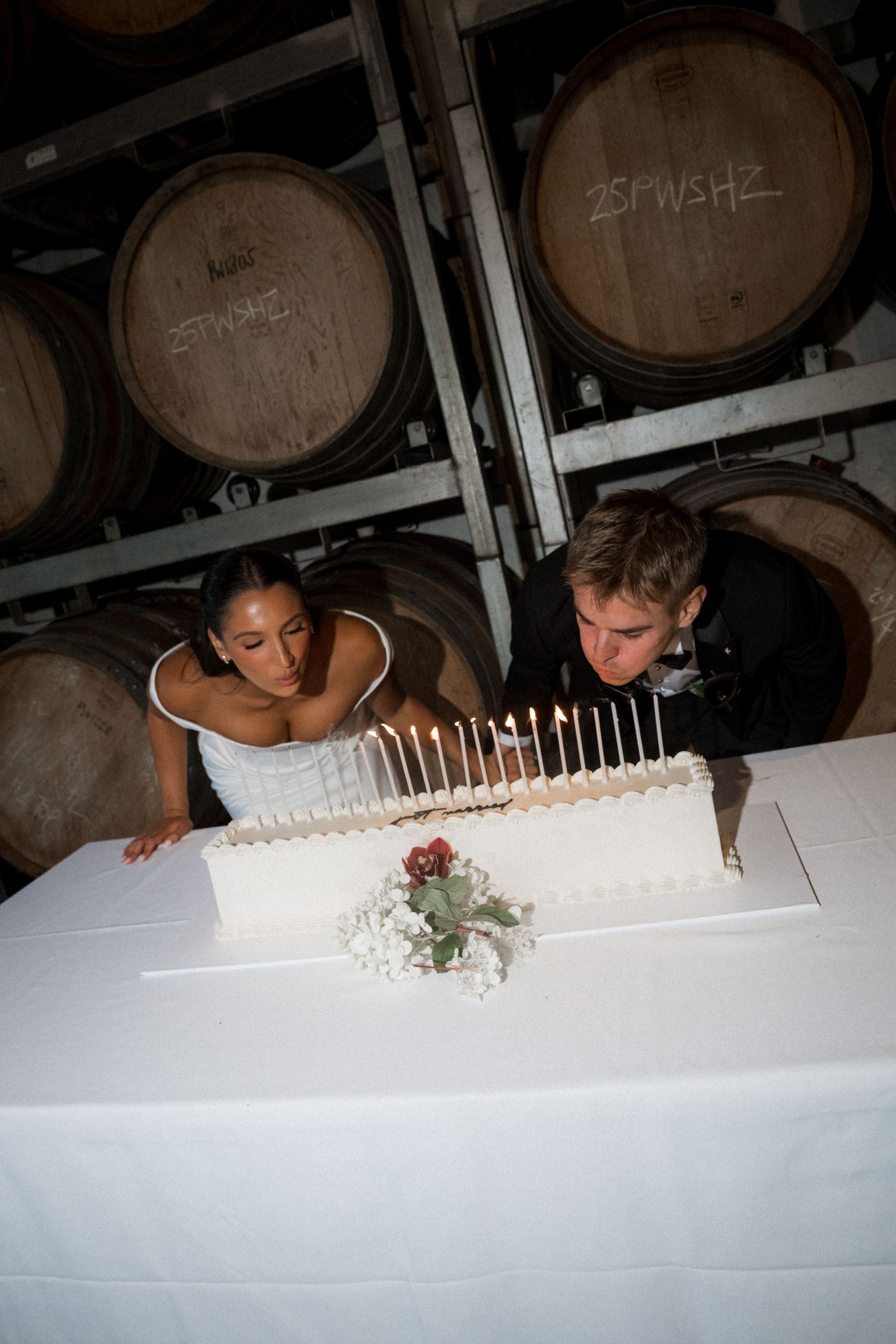 Couple signing their wedding guest book together at a rustic venue with wine barrels in the background.