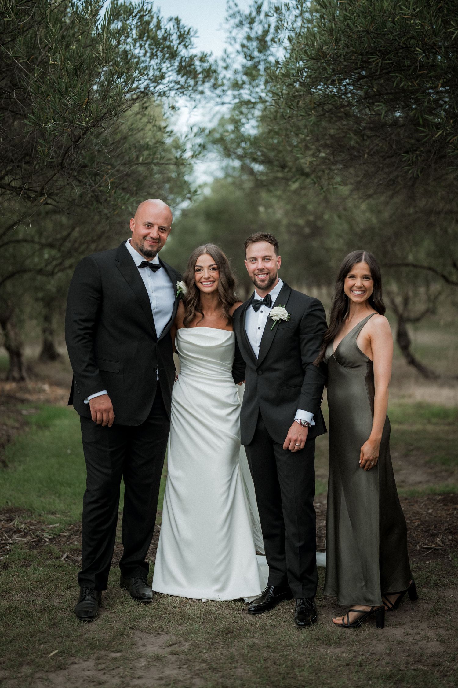 Wedding party poses together outdoors among olive trees wearing formal attire.