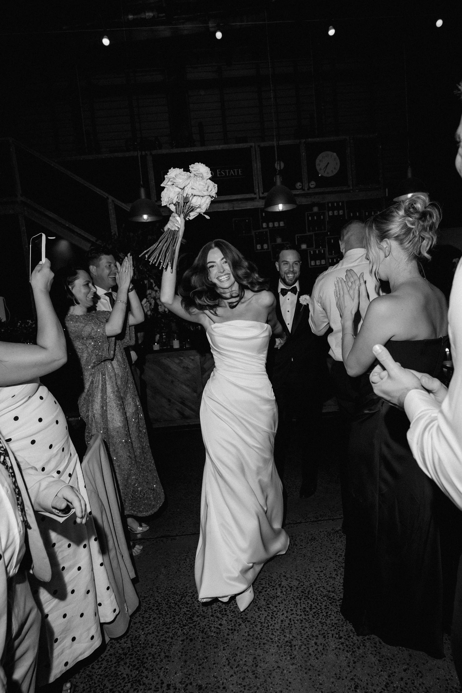 A bride in a white dress dances joyfully at a wedding reception holding her bouquet high in the air.