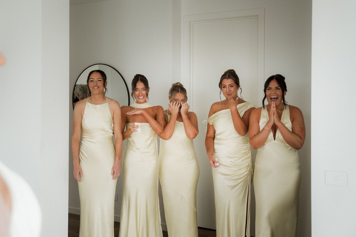 Five bridesmaids in elegant cream-colored dresses pose together in a bright room.
