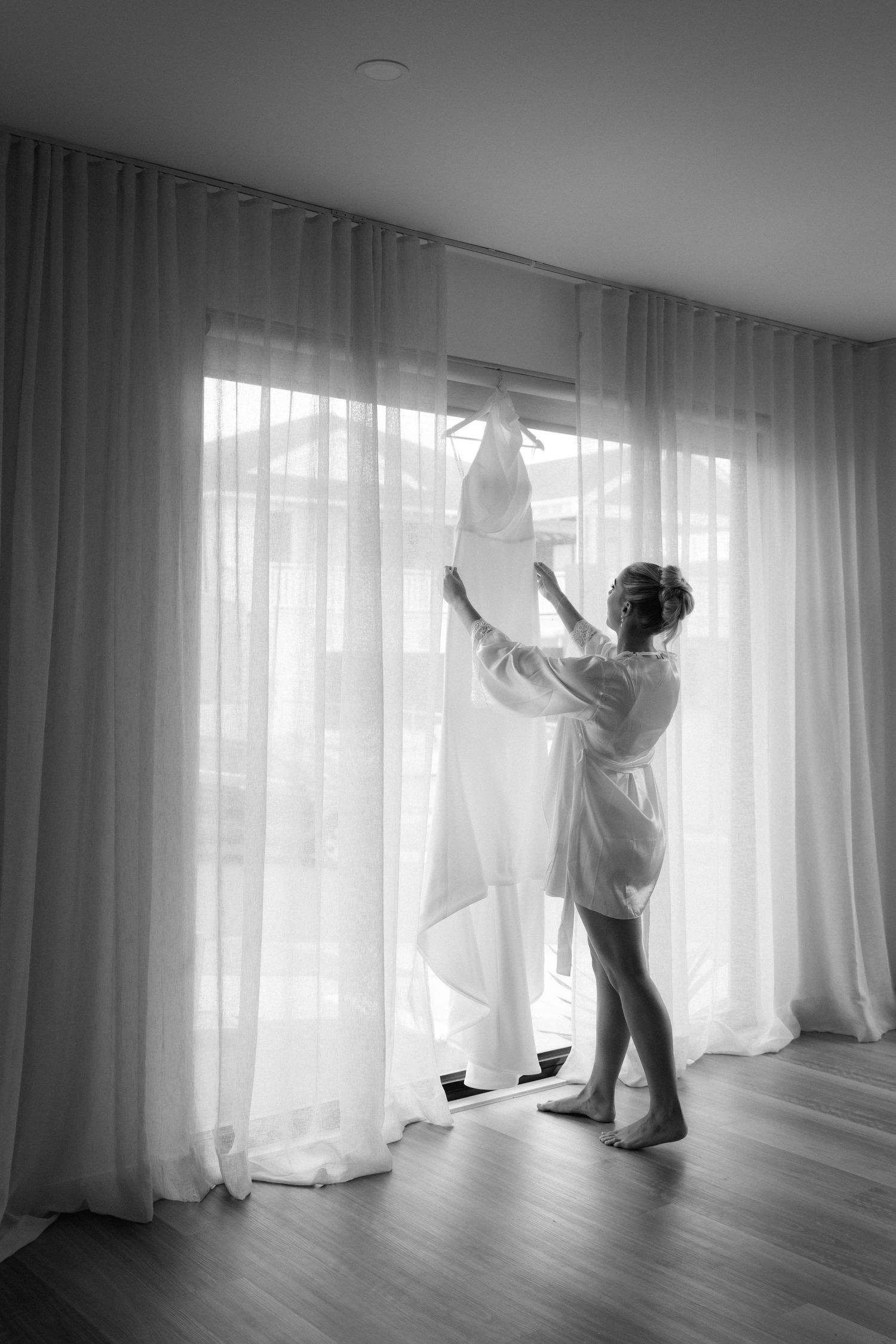 Person adjusting white curtains in a bright, minimalist room with wooden floors.
