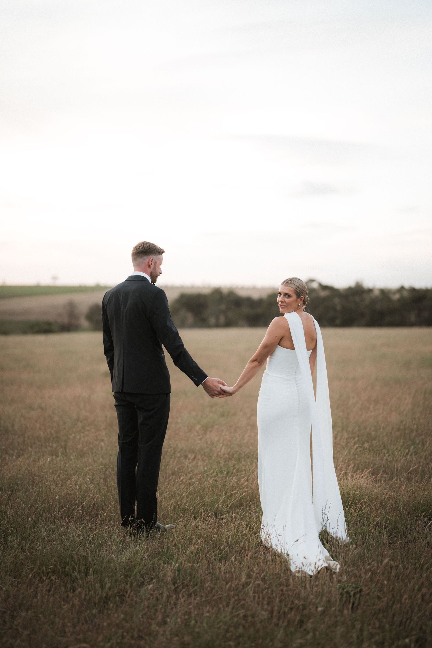 Couple walks hand in hand through rustic field at golden hour.