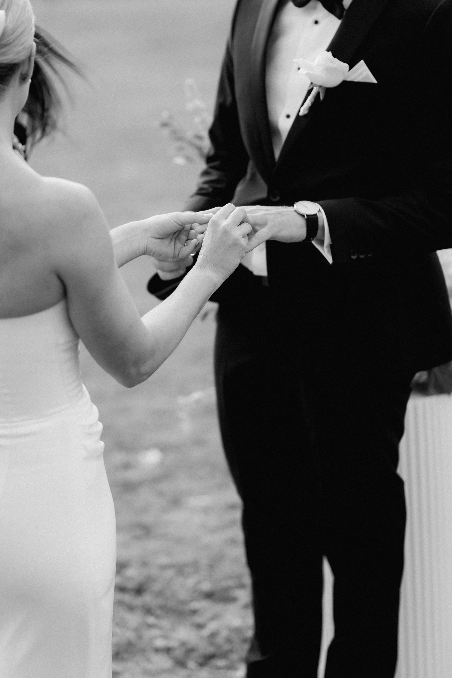A couple in formal wedding attire exchange rings during an outdoor ceremony.